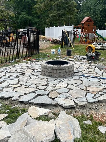 Stone fire pit on a flagstone patio in a backyard, with a playset and fenced yard in the background.