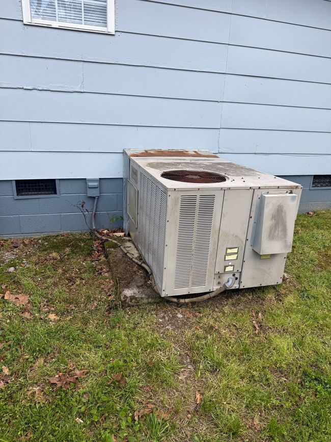 Outdoor AC unit beside a light blue house, sitting on grass and leaves.