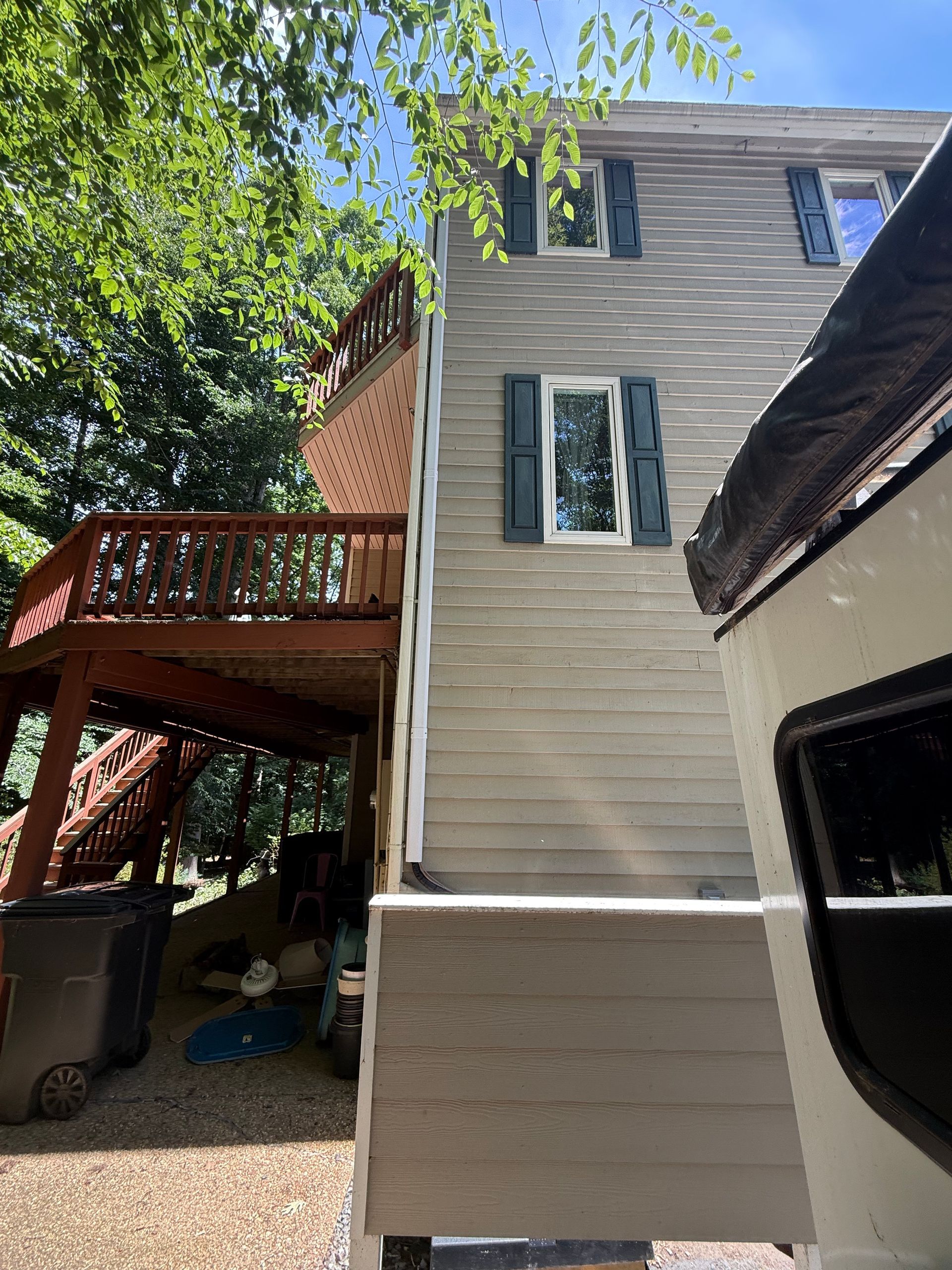Side view of a beige house with a wooden deck, stairs, and a gravel yard under bright sunlight.
