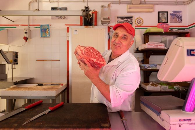 Una persona con un berretto rosso tiene in mano un grosso taglio di carne in una macelleria.