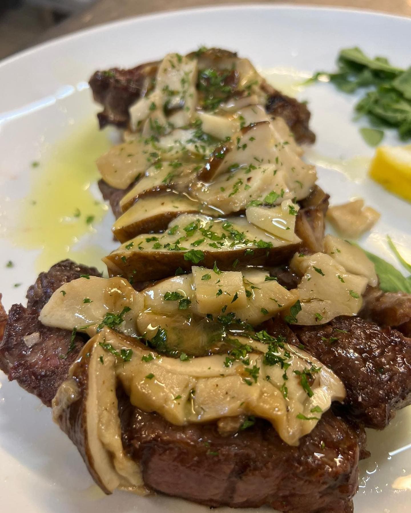 Bistecca alla griglia con funghi ed erbe aromatiche su un piatto bianco con limone e verdure.