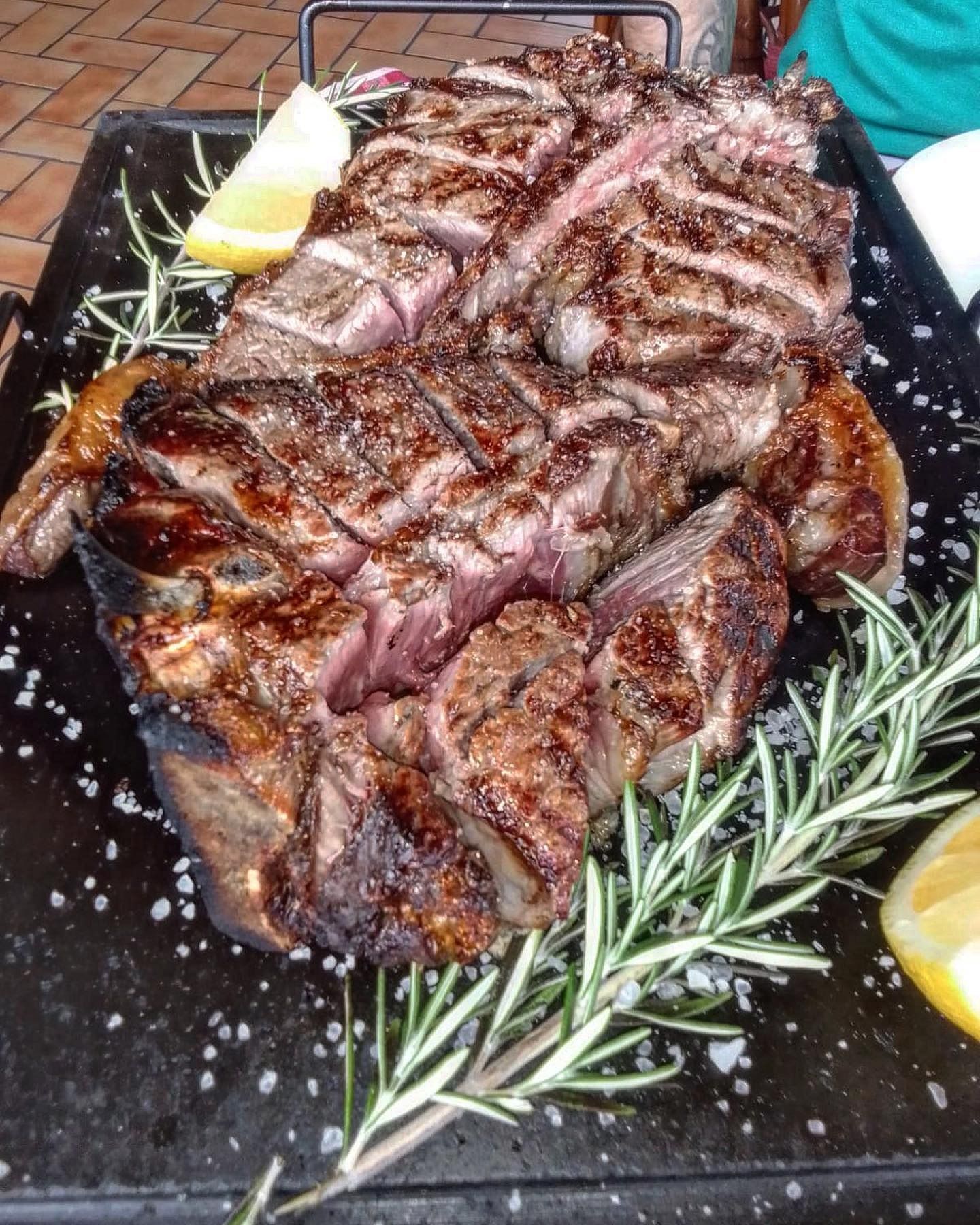 Bistecca alla griglia su un piatto con rametti di rosmarino e spicchi di limone su un tavolo di mattoni.