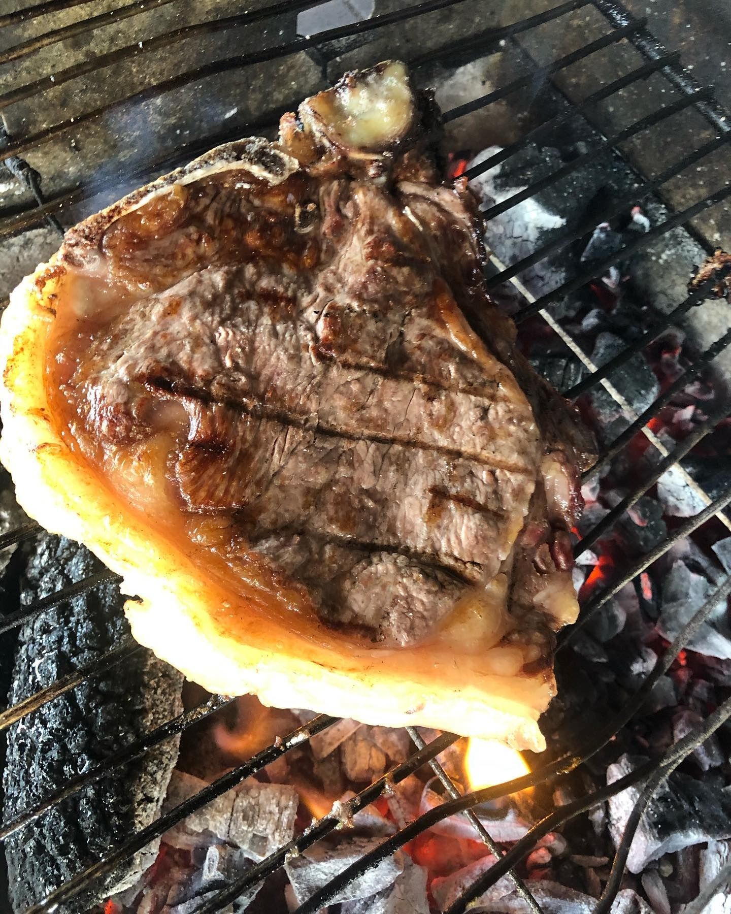 Bistecca cotta alla griglia su braci ardenti su un barbecue, con segni di bruciatura e grasso fuso.