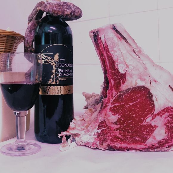 Bottiglia di vino e bicchiere accanto a una grande bistecca di controfiletto cruda su una superficie chiara.