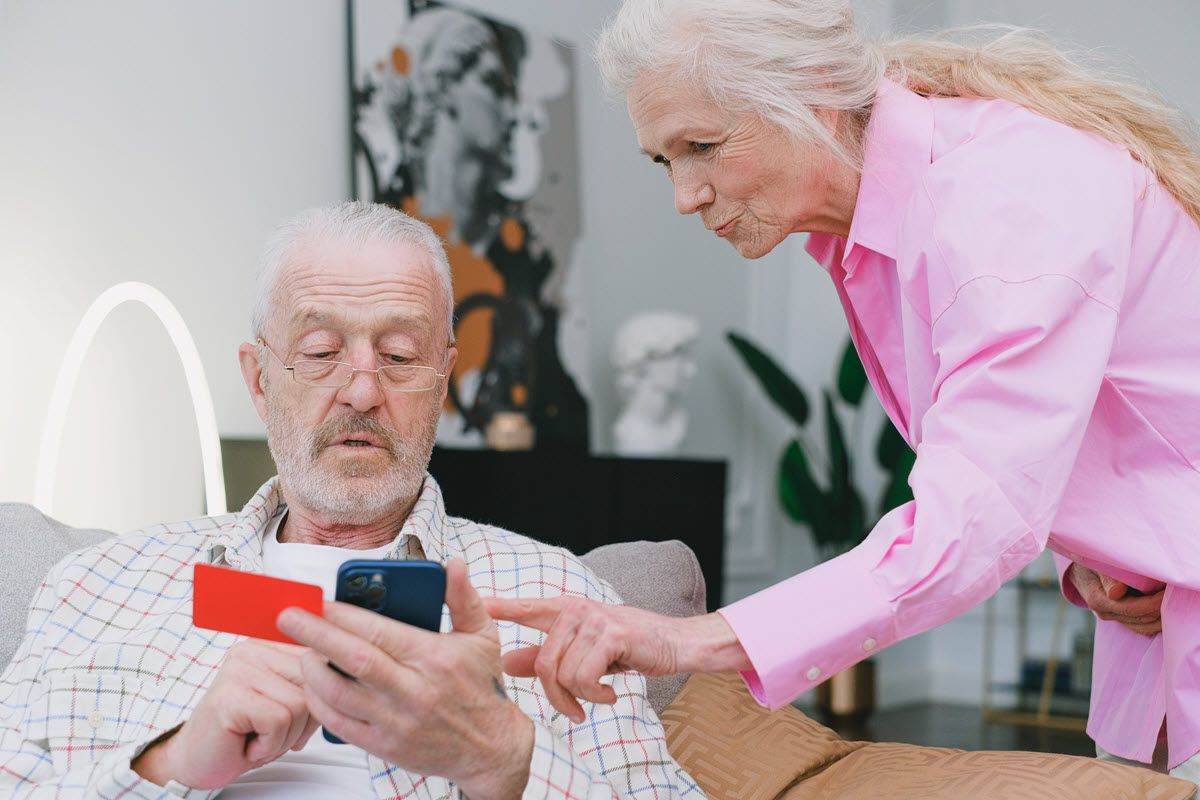 Elderly man on a couch holding a phone while an elderly woman in pink leans over to help him