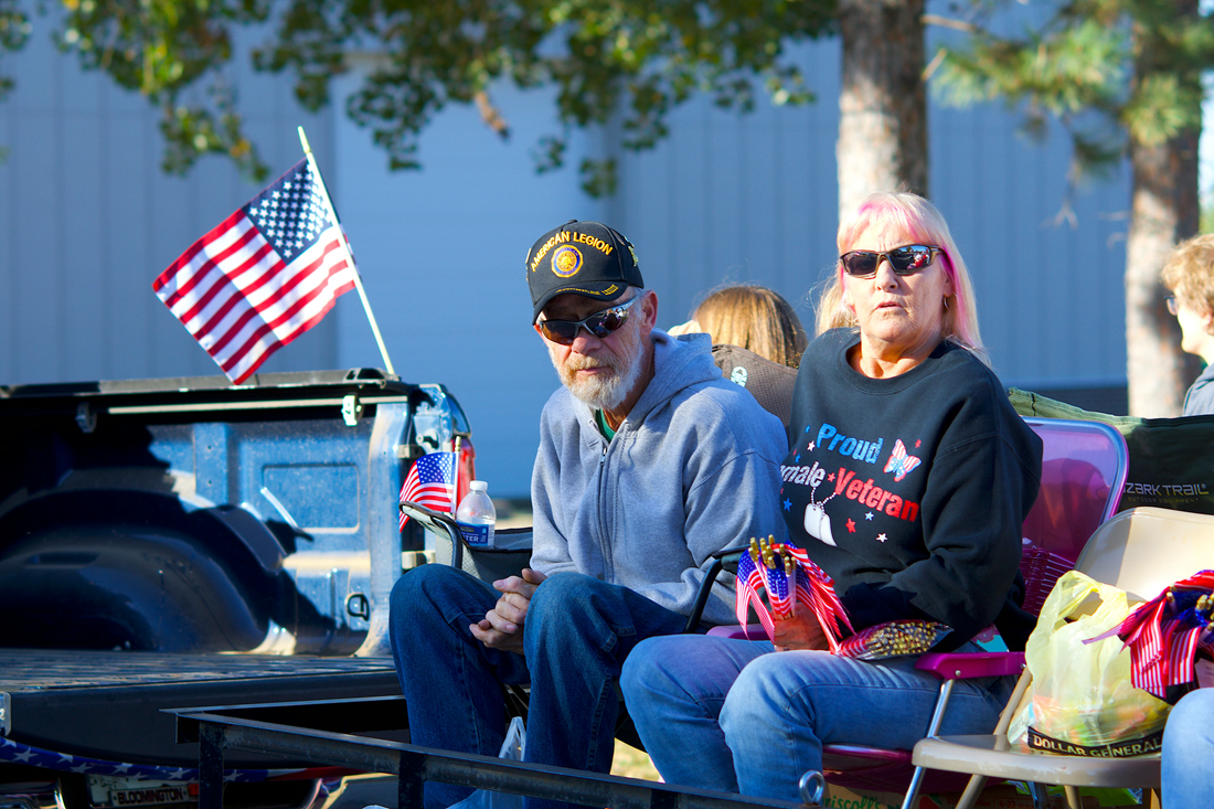 Two people seated outdoors beside a small American flag, with bags and a blue vehicle in the background