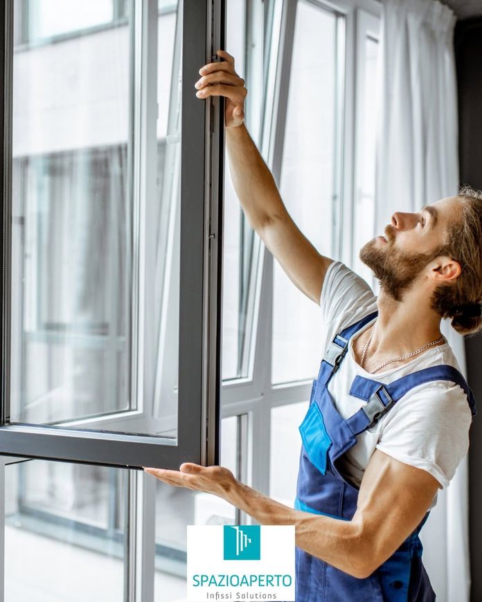 Un operaio in tuta blu sta installando un telaio di finestra all'interno di un edificio, allungando una mano verso l'alto mentre tiene fermo il telaio.