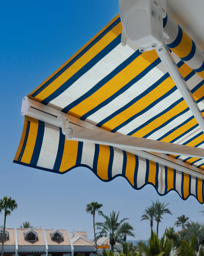 Tenda a strisce gialle, blu e bianche contro un cielo luminoso, con palme e tetti sottostanti.