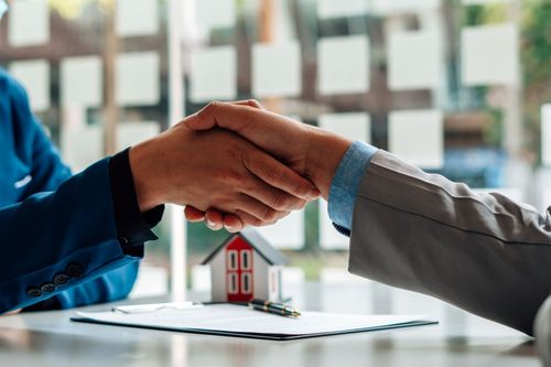 Two people shaking hands over a table with a small house model and paperwork