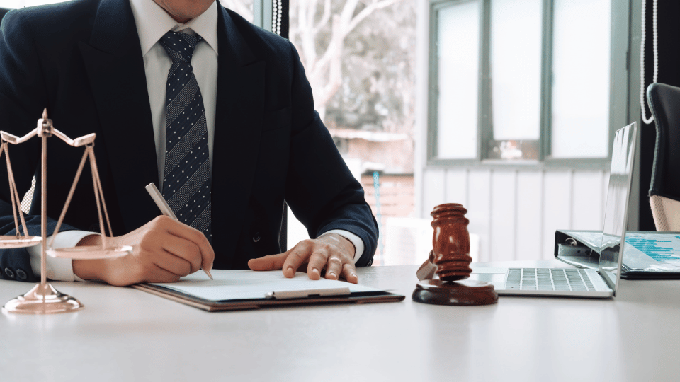 Lawyer in a dark suit writing at a desk with scales of justice, gavel, and laptop nearby