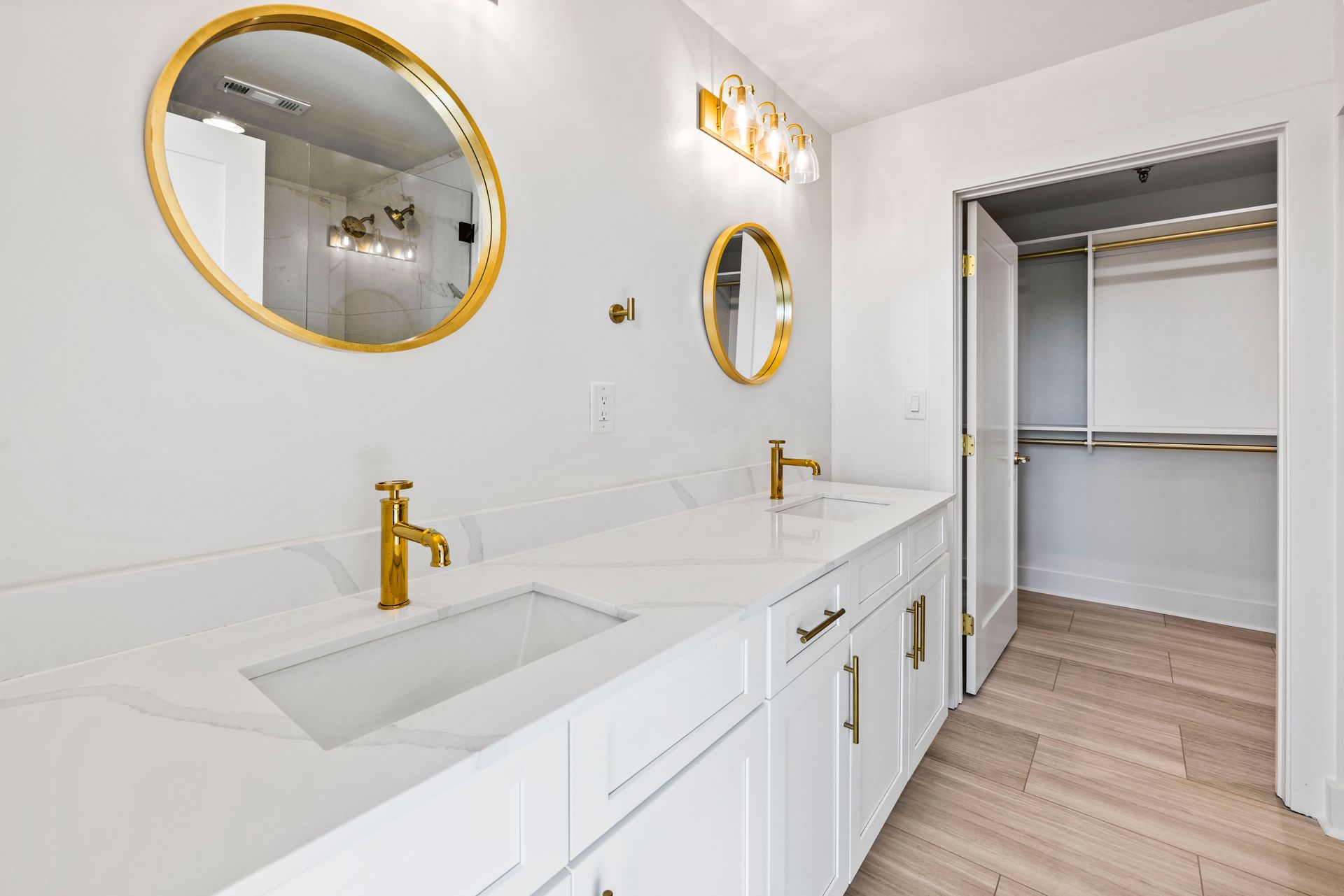 Bright bathroom with white cabinets, gold fixtures, and two round mirrors. Closet on right.