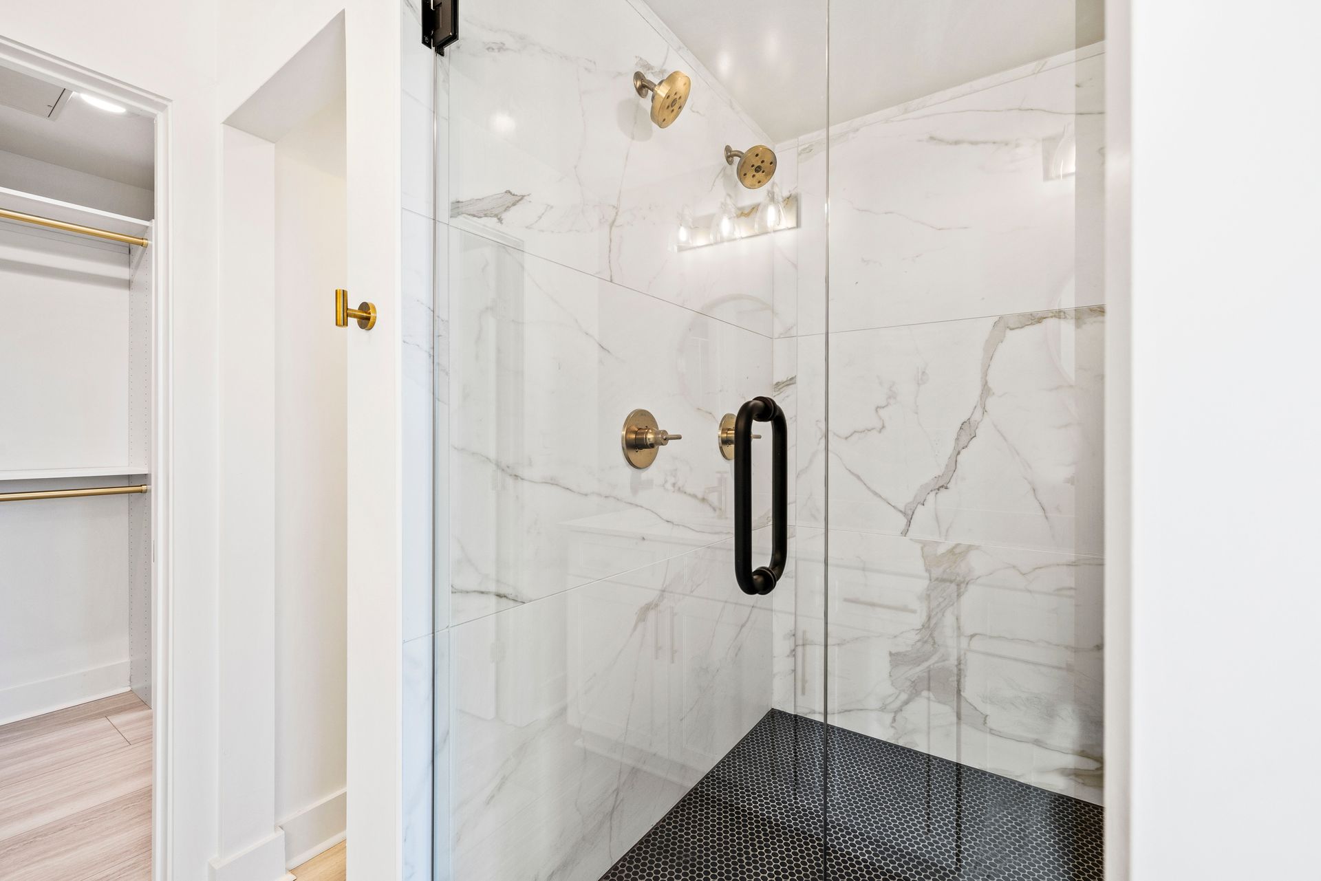 Marble-tiled shower with a glass door and gold fixtures. Black and white mosaic tile floor. Adjacent is a walk-in closet.