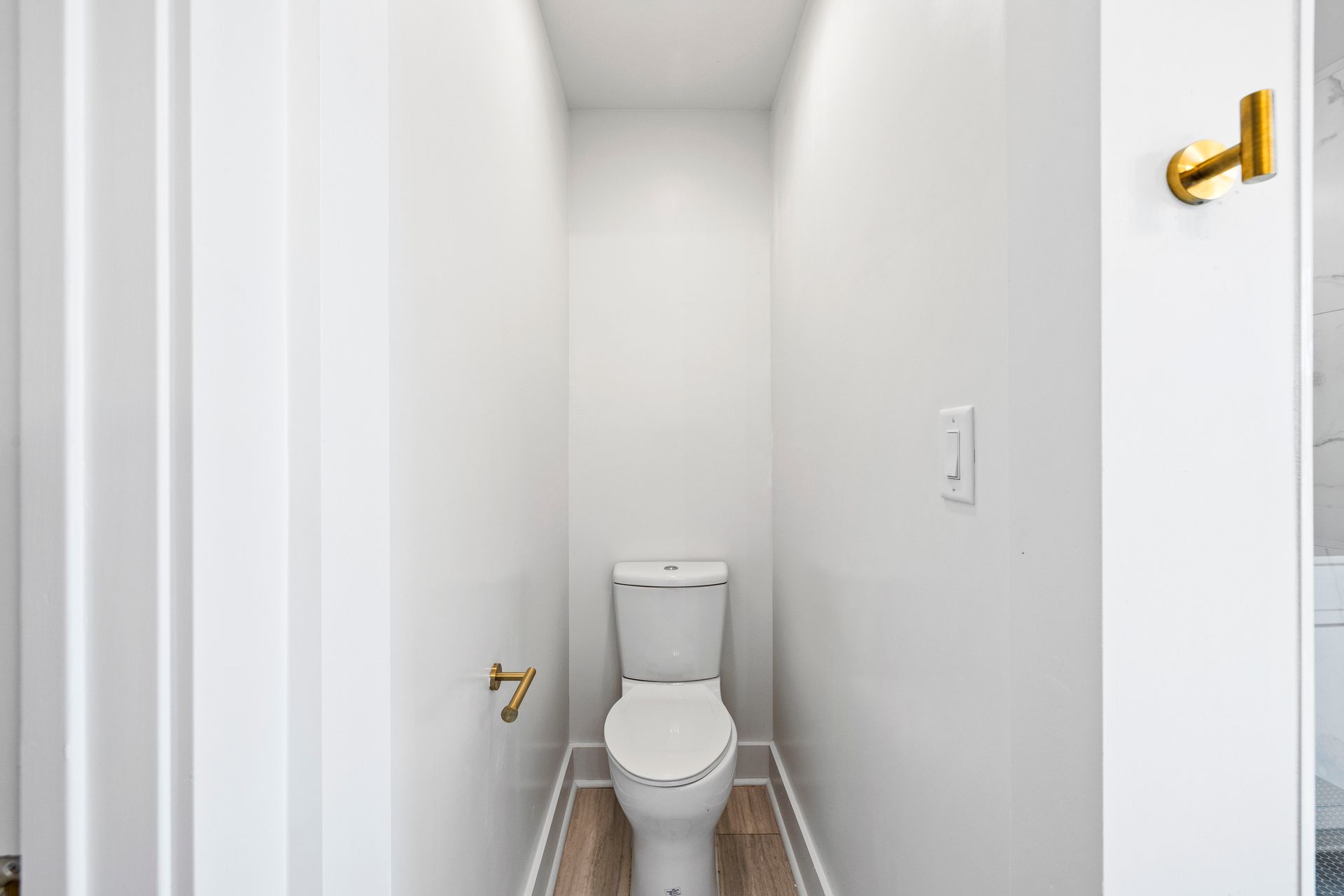 Small, narrow, white bathroom with toilet and gold fixtures. Wooden floor, light switch on wall.
