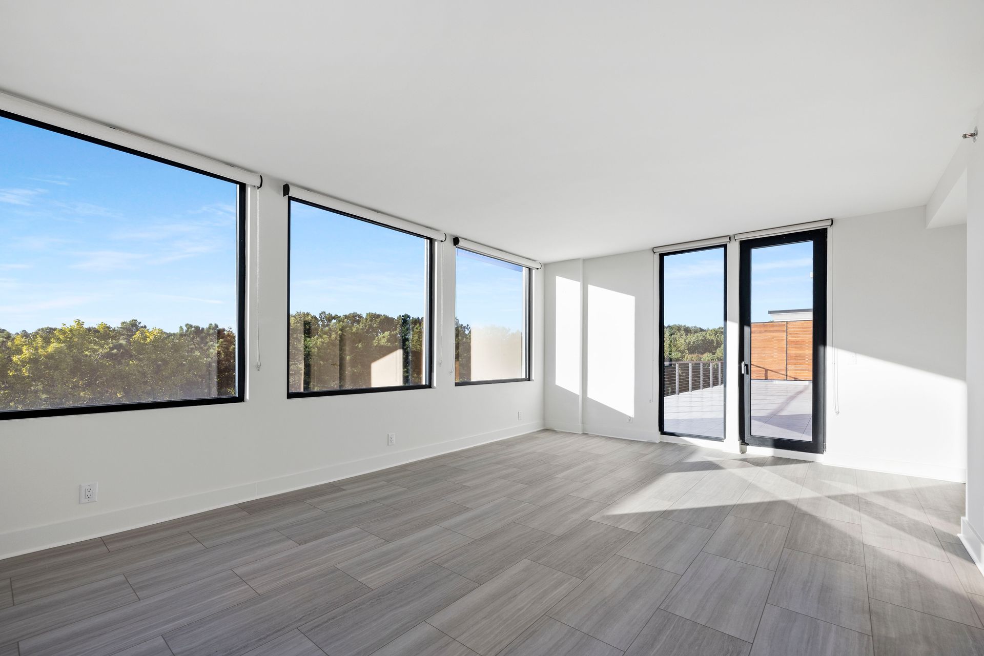 Empty room with large windows, light gray floors, white walls, and a view of greenery outside.