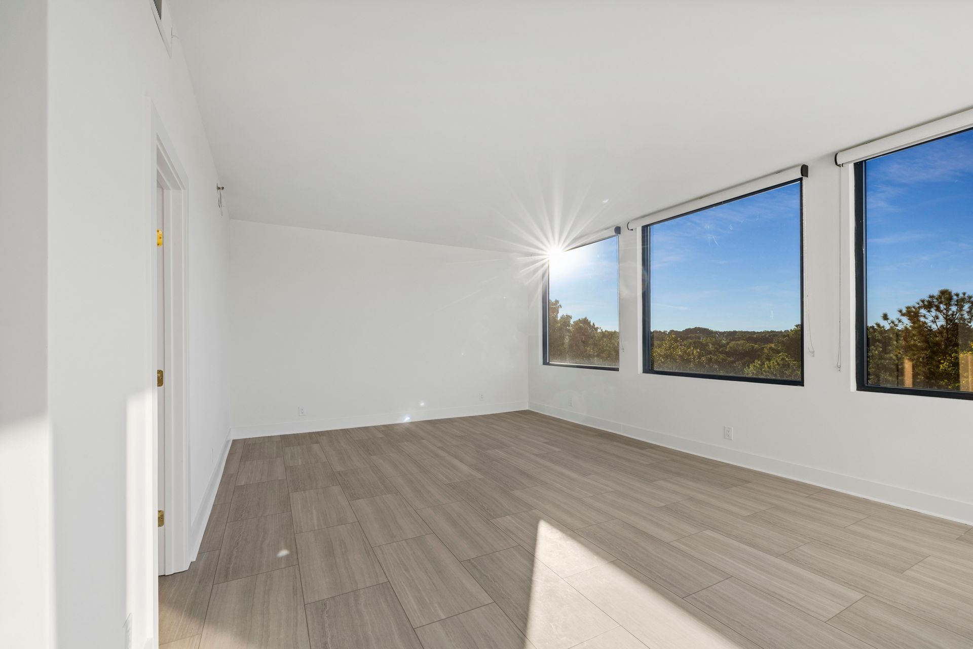 Empty room with large windows overlooking a sunny outdoor view and light wood flooring.