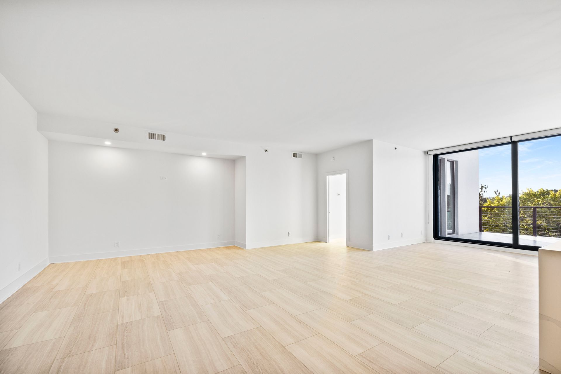 Empty, bright living room with blonde wood floors and large windows overlooking greenery.