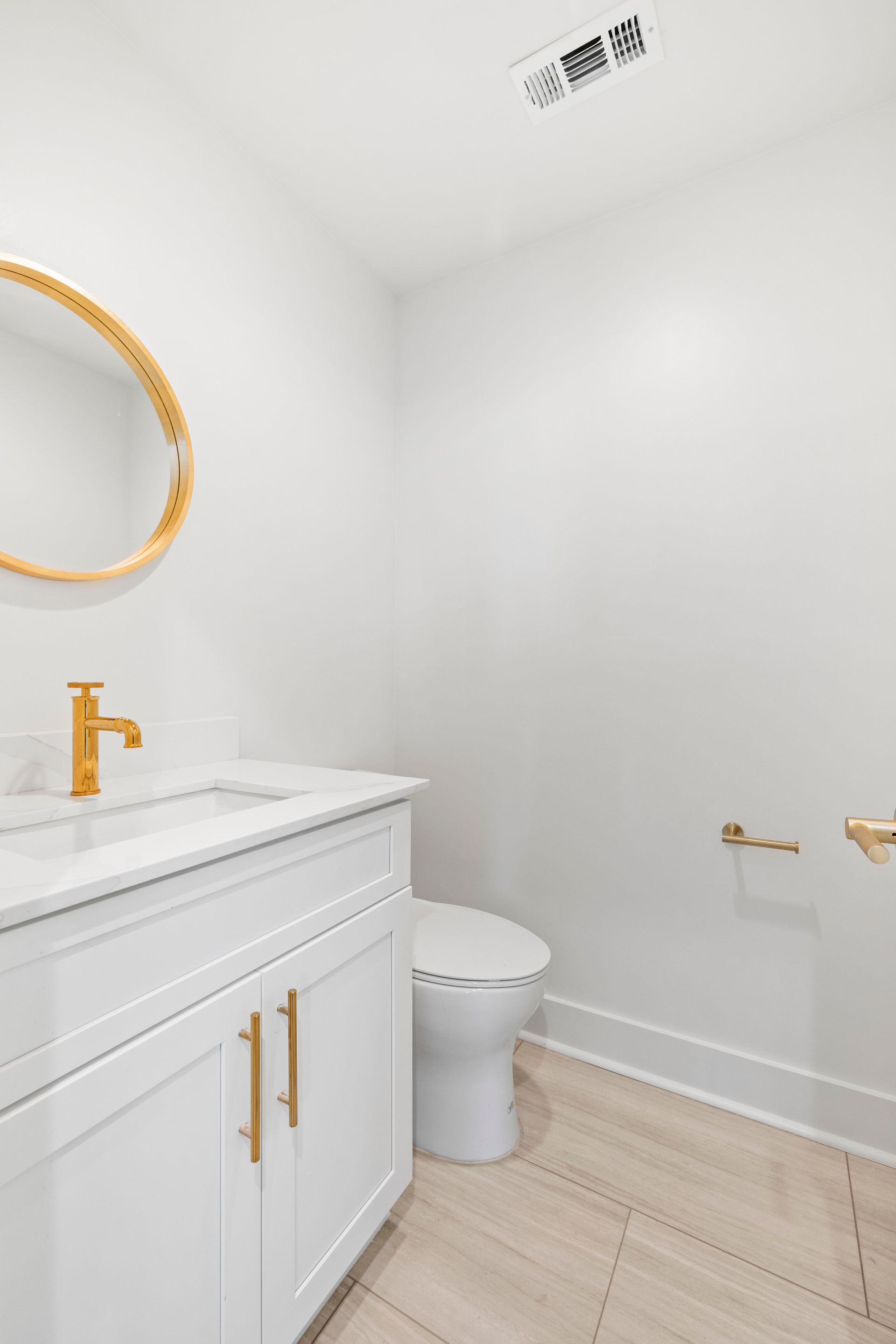 White bathroom with gold fixtures. Vanity, toilet, and a round mirror.
