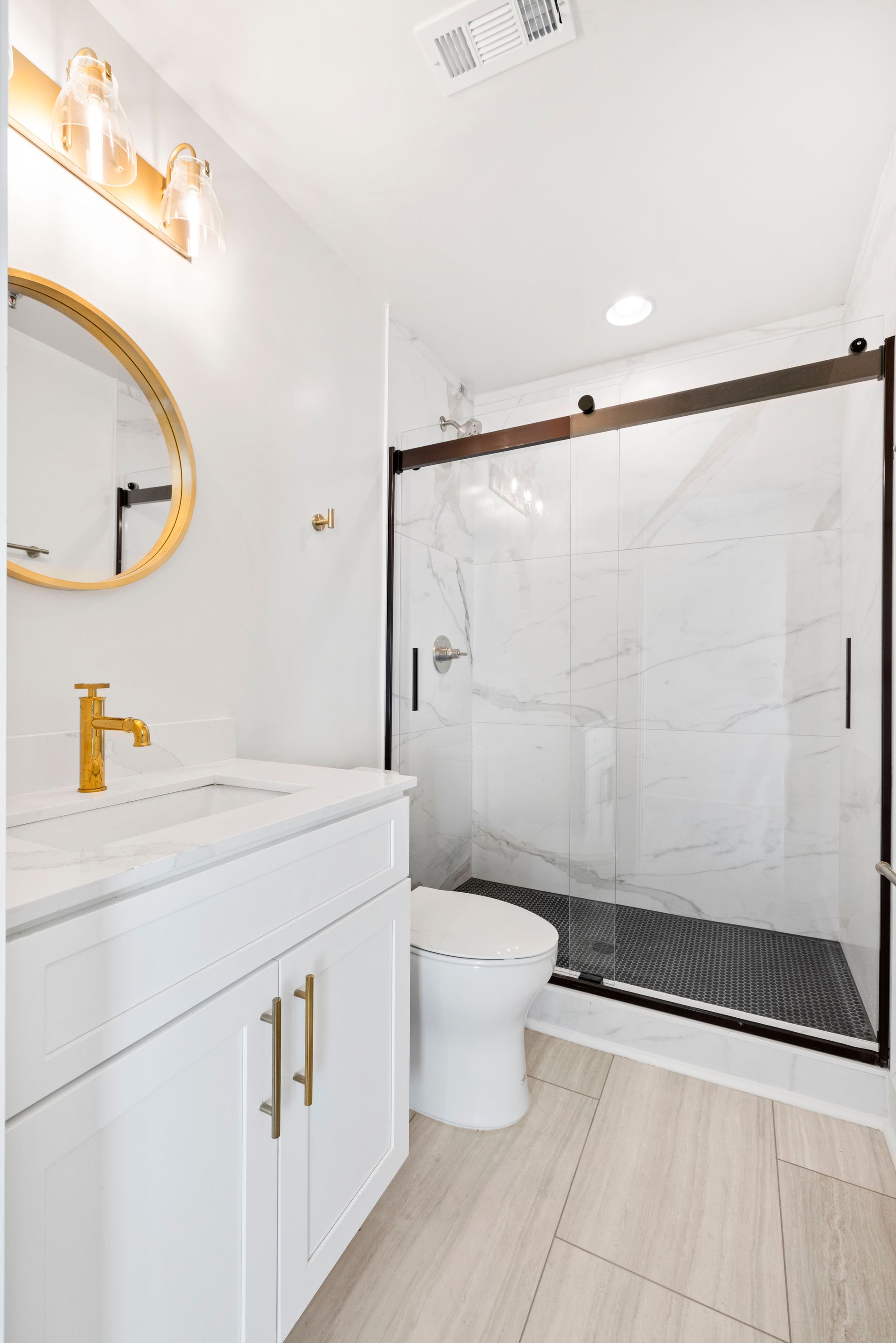 Modern white bathroom with gold accents, marble shower, and sliding glass door.