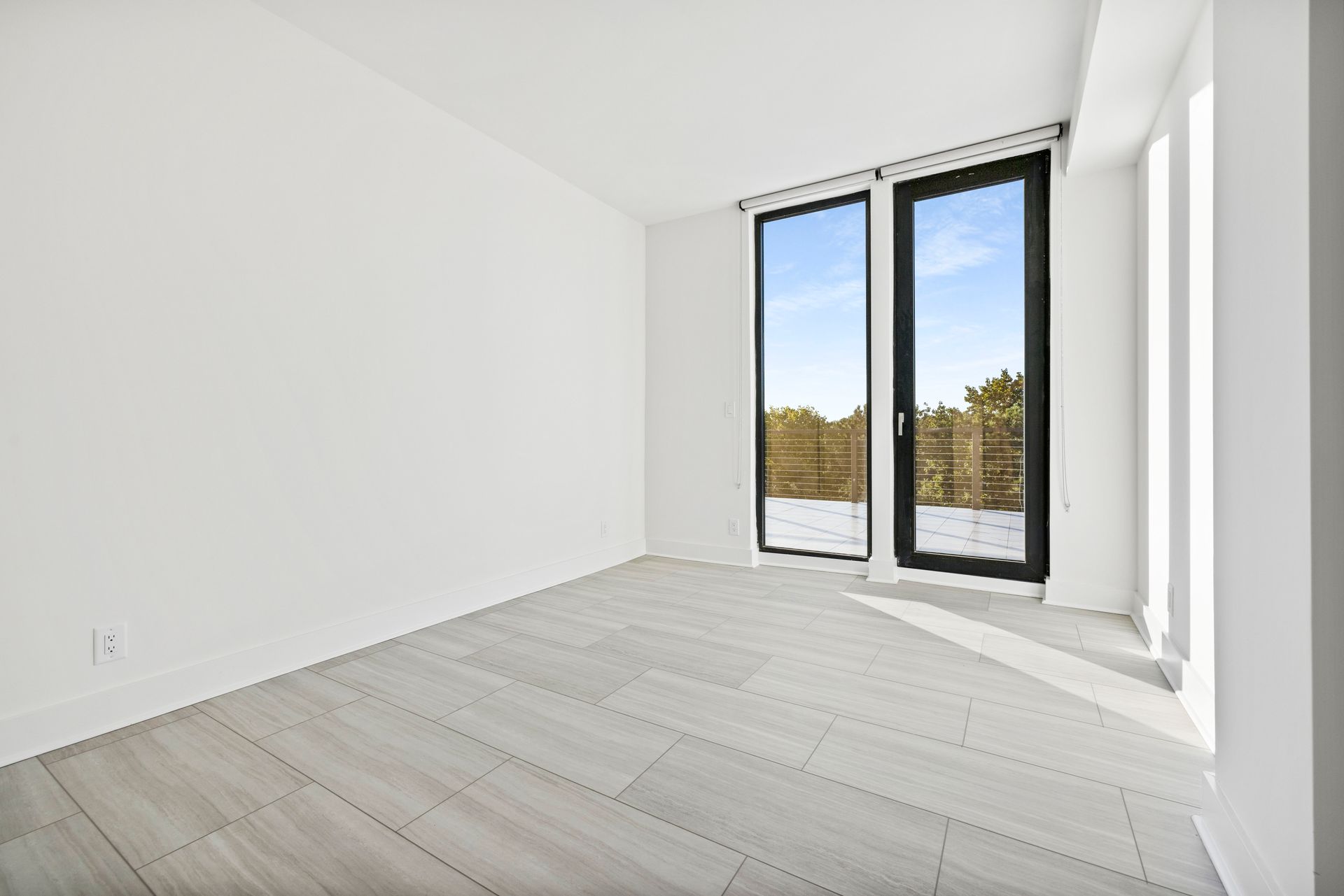 Empty room with gray wood-look floor, white walls, and a large window with a view of trees and sky.