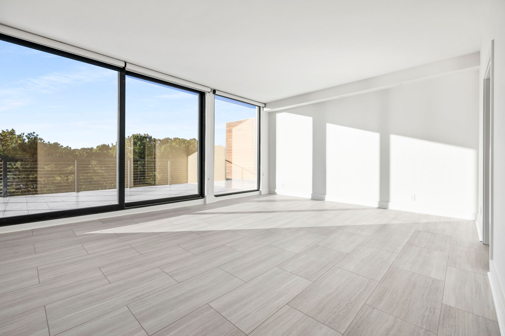 Empty, sunlit room with large windows overlooking a tree-lined area. Light gray floor, white walls.