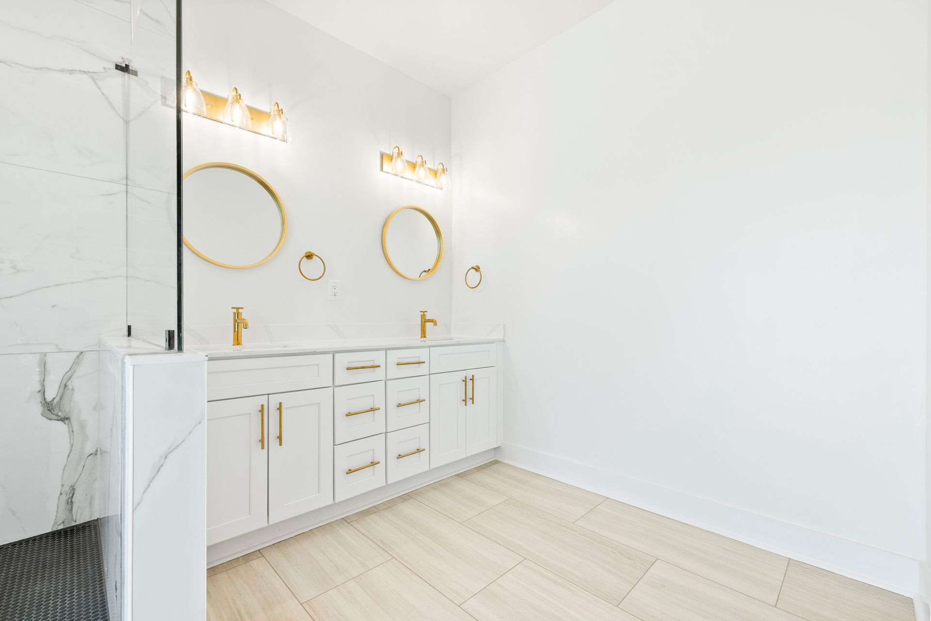 White bathroom with gold fixtures: double vanity, round mirrors, sconces, marble shower, and light wood flooring.