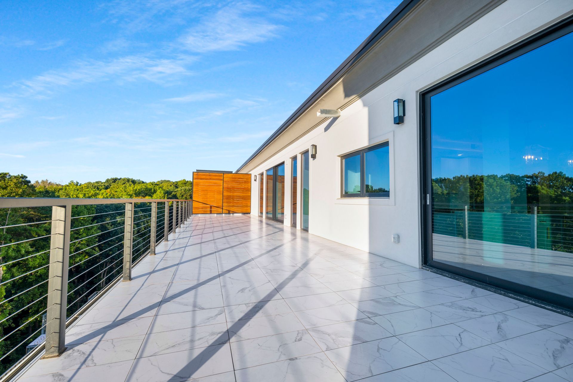 Rooftop patio with gray floor, cable railing, and large windows reflecting blue sky and trees.