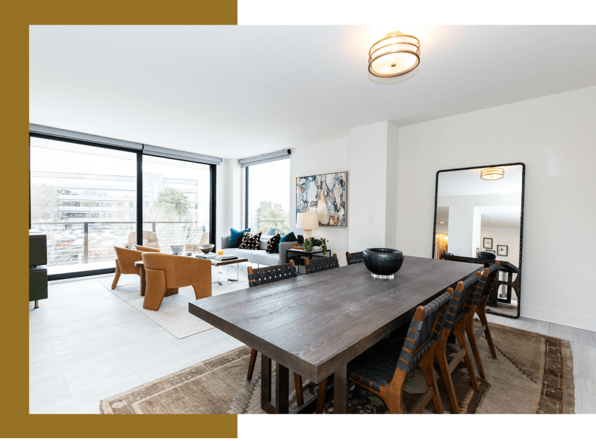 Modern dining room with large table, chairs, rug, and full-length mirror. Living area has a view of the outdoors.