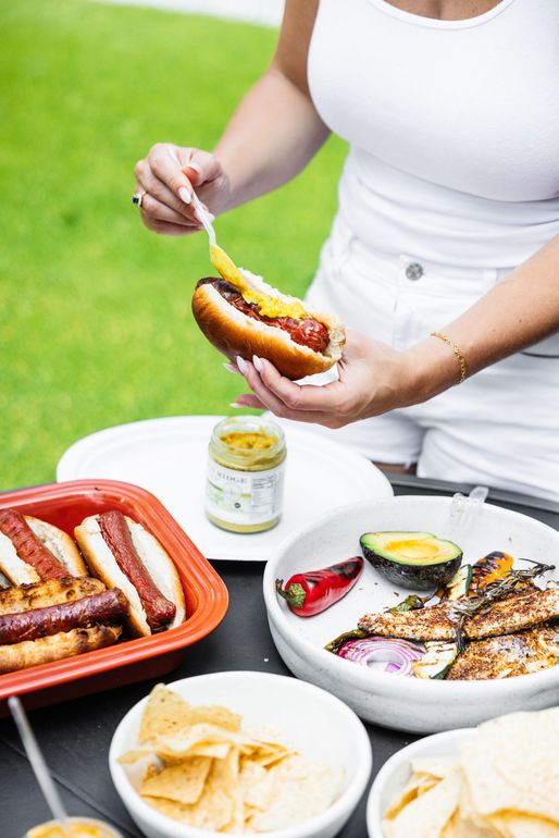 A woman is dipping a hot dog in mustard at a picnic table.
