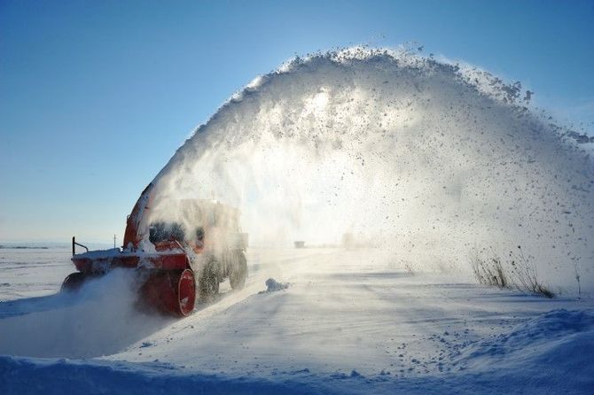 Red snowplow throwing a huge arc of snow across a snowy landscape under a blue sky