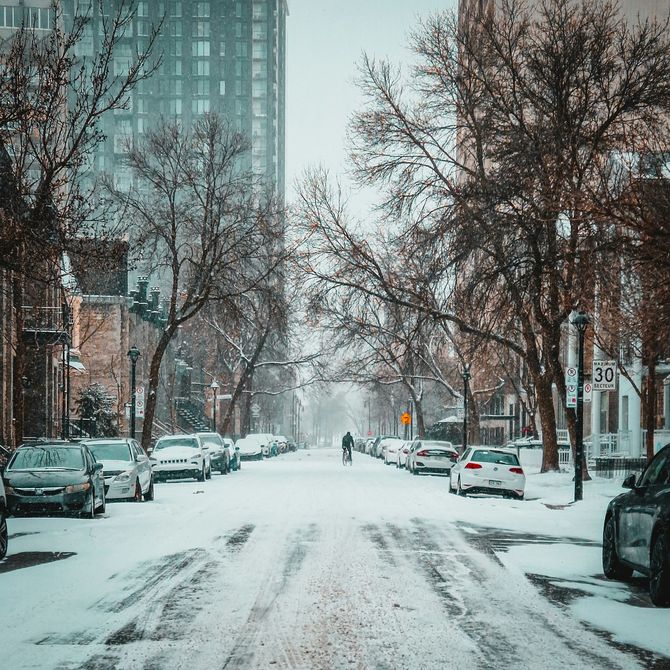 Snow-covered city street with parked cars, leafless trees, and a lone pedestrian in the distance.