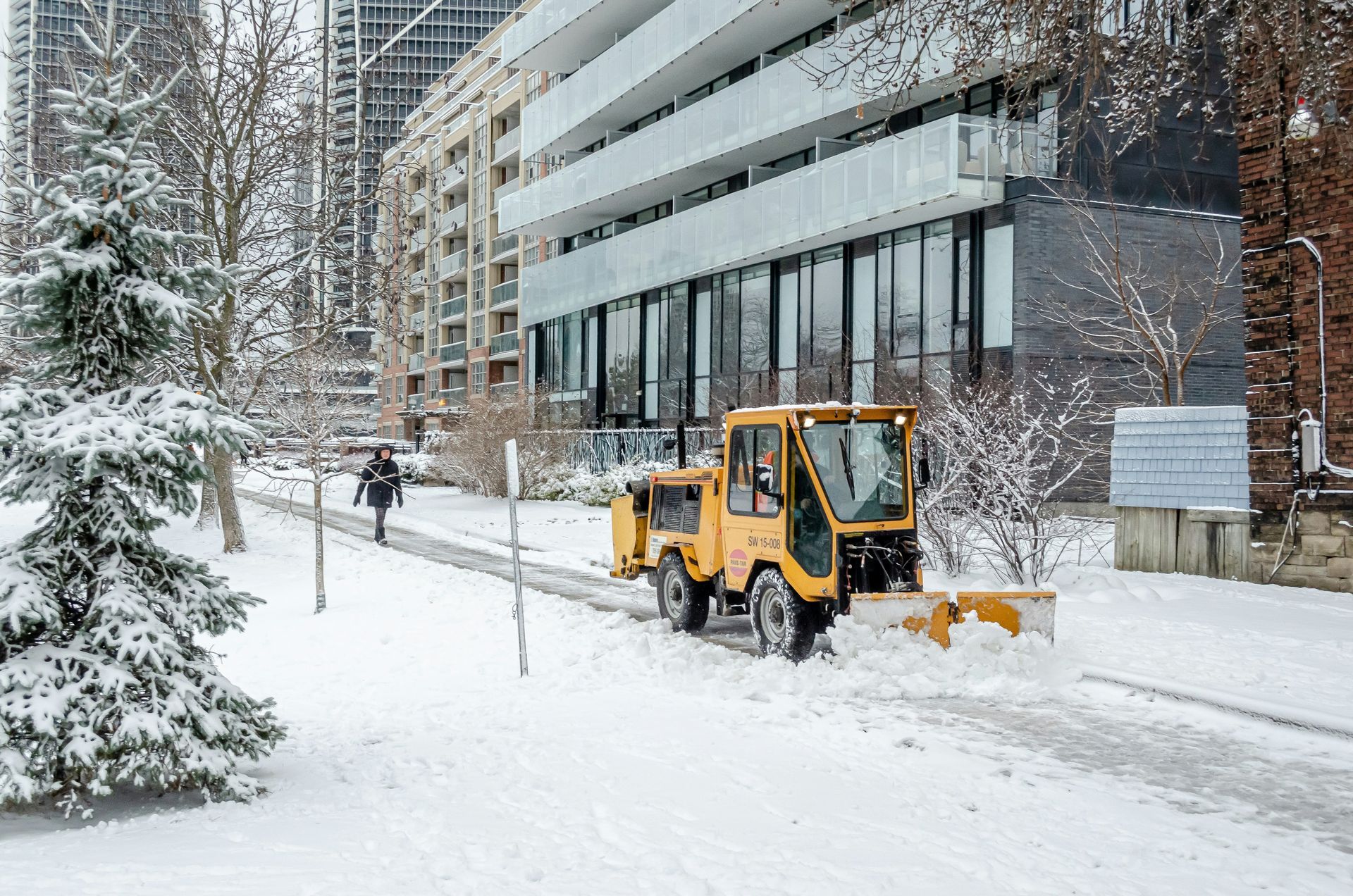 Snow-covered city sidewalk with a yellow snowplow clearing path beside a modern building