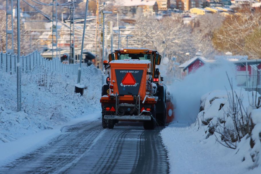 Orange snowplow clearing a snowy road lined with trees and houses