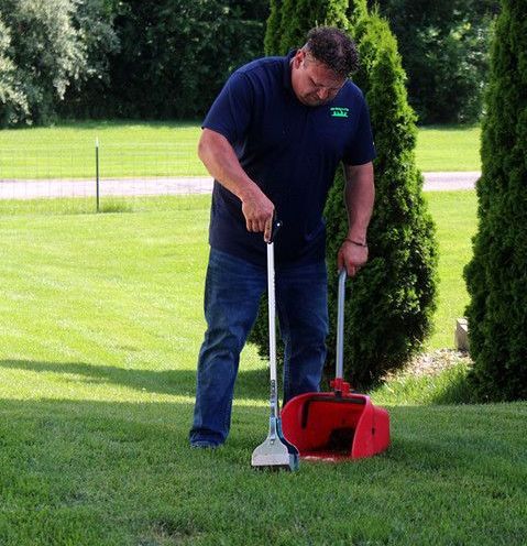 A man is standing in a lush green field holding a red scoop and shovel.