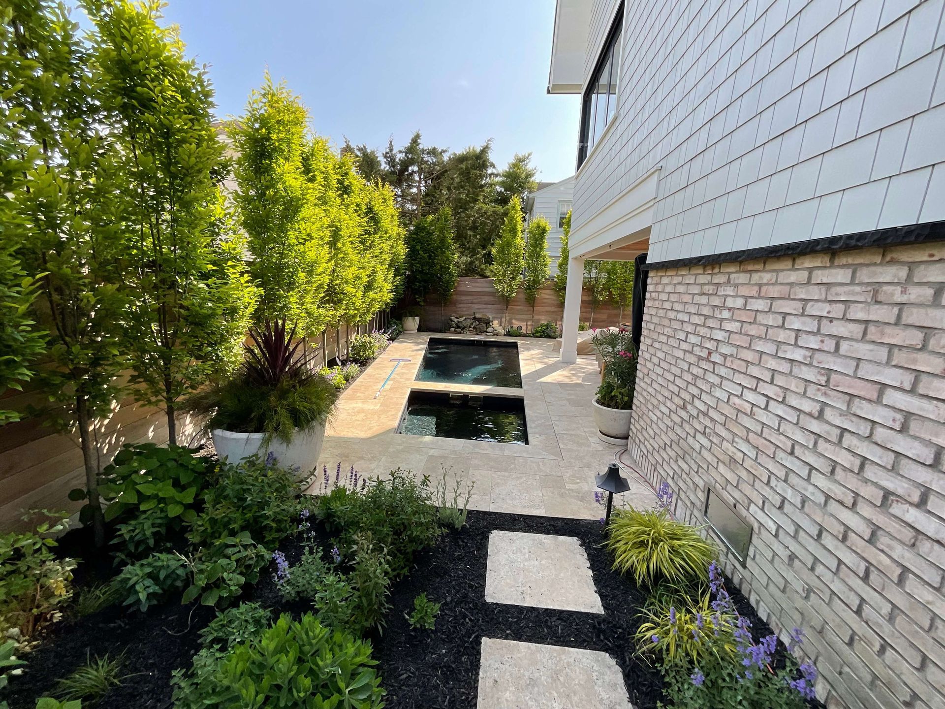 Modern backyard patio with stone path, raised water feature, brick house wall, and green trees along the garden.