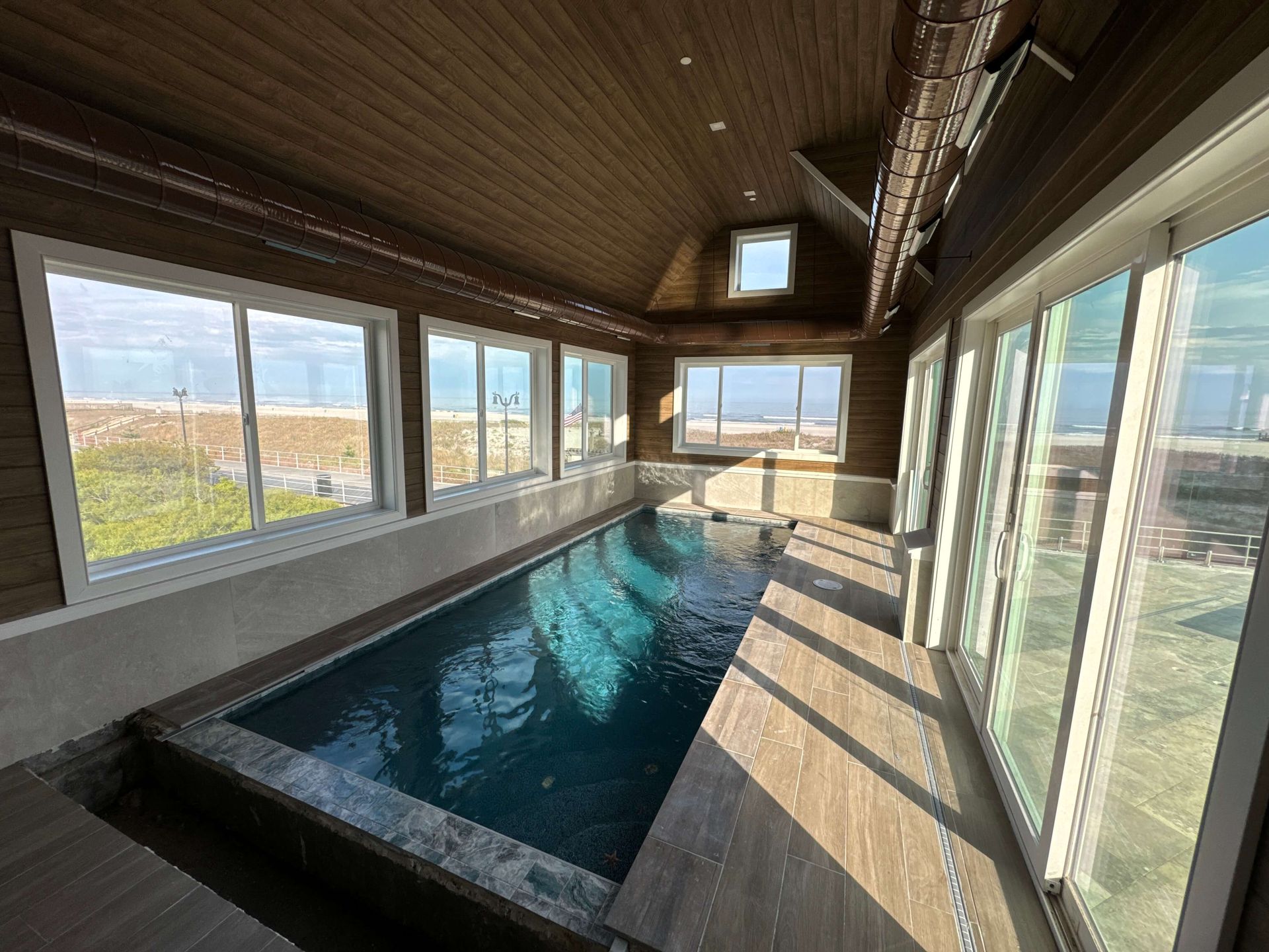 Indoor lap pool with large windows, sunlight, and wood-paneled ceiling
