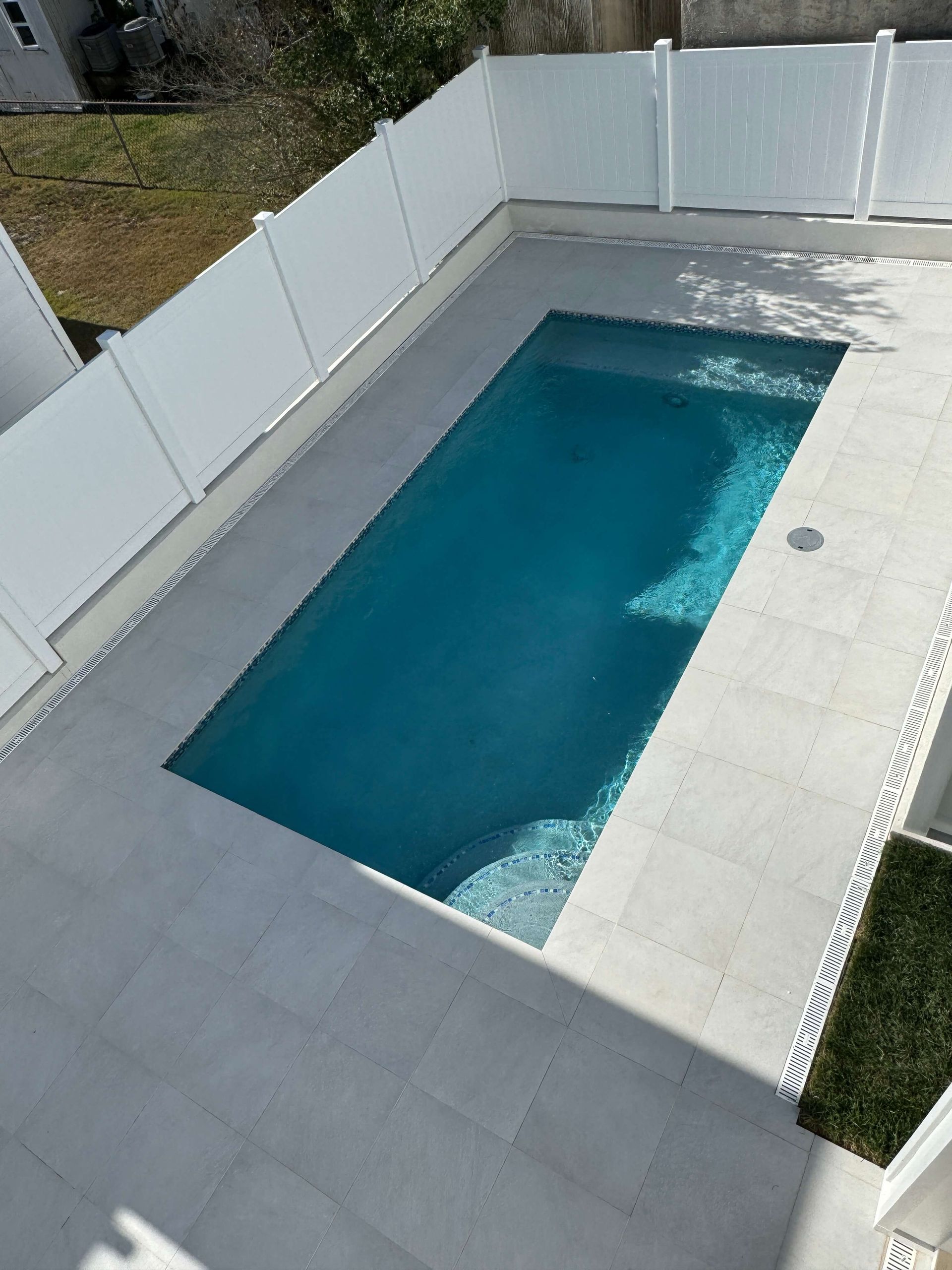 A backyard swimming pool with blue water, surrounded by gray paving and white privacy fencing.