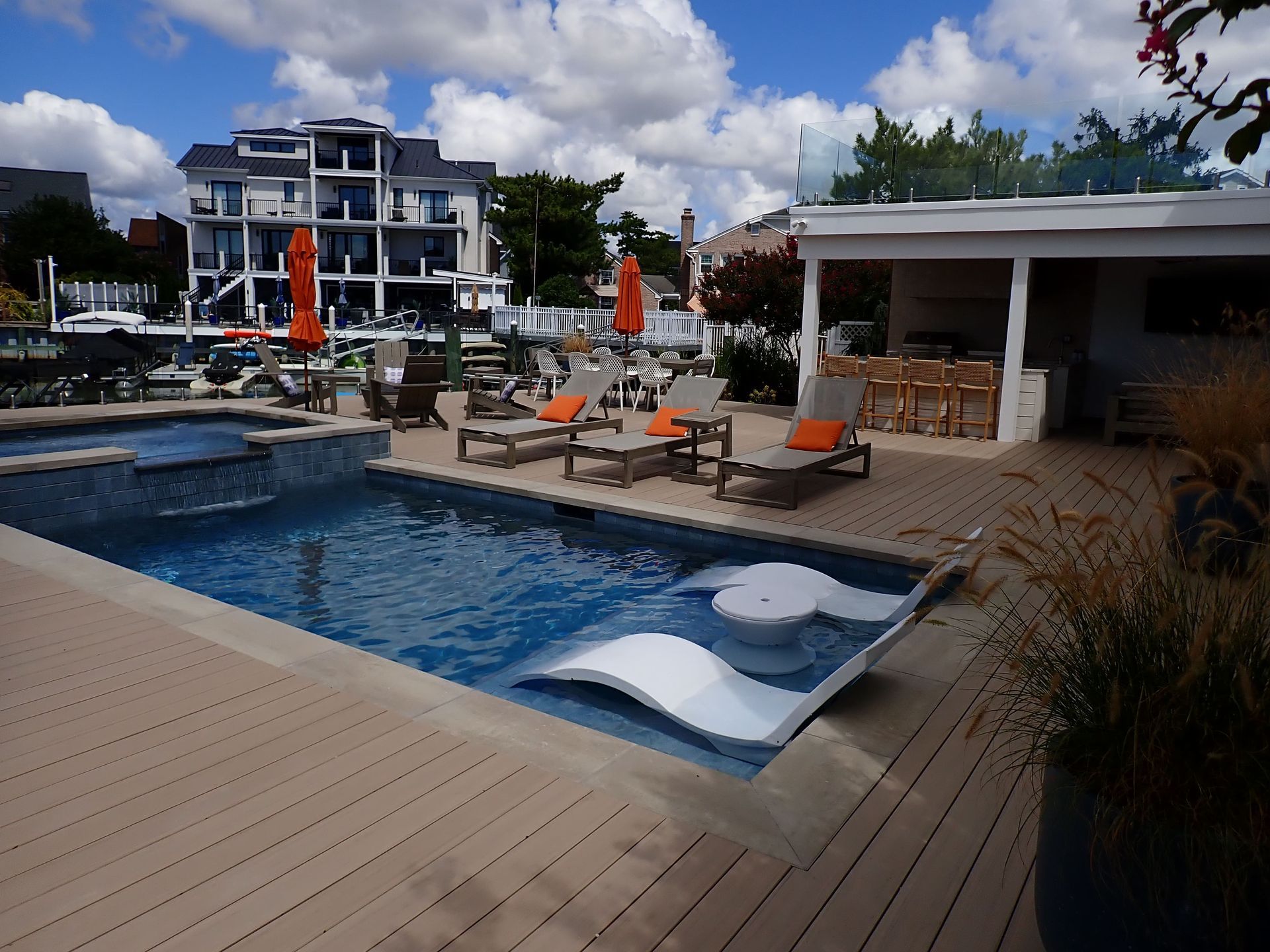 Resort pool with lounge chairs, white cabana, and modern buildings under a partly cloudy sky