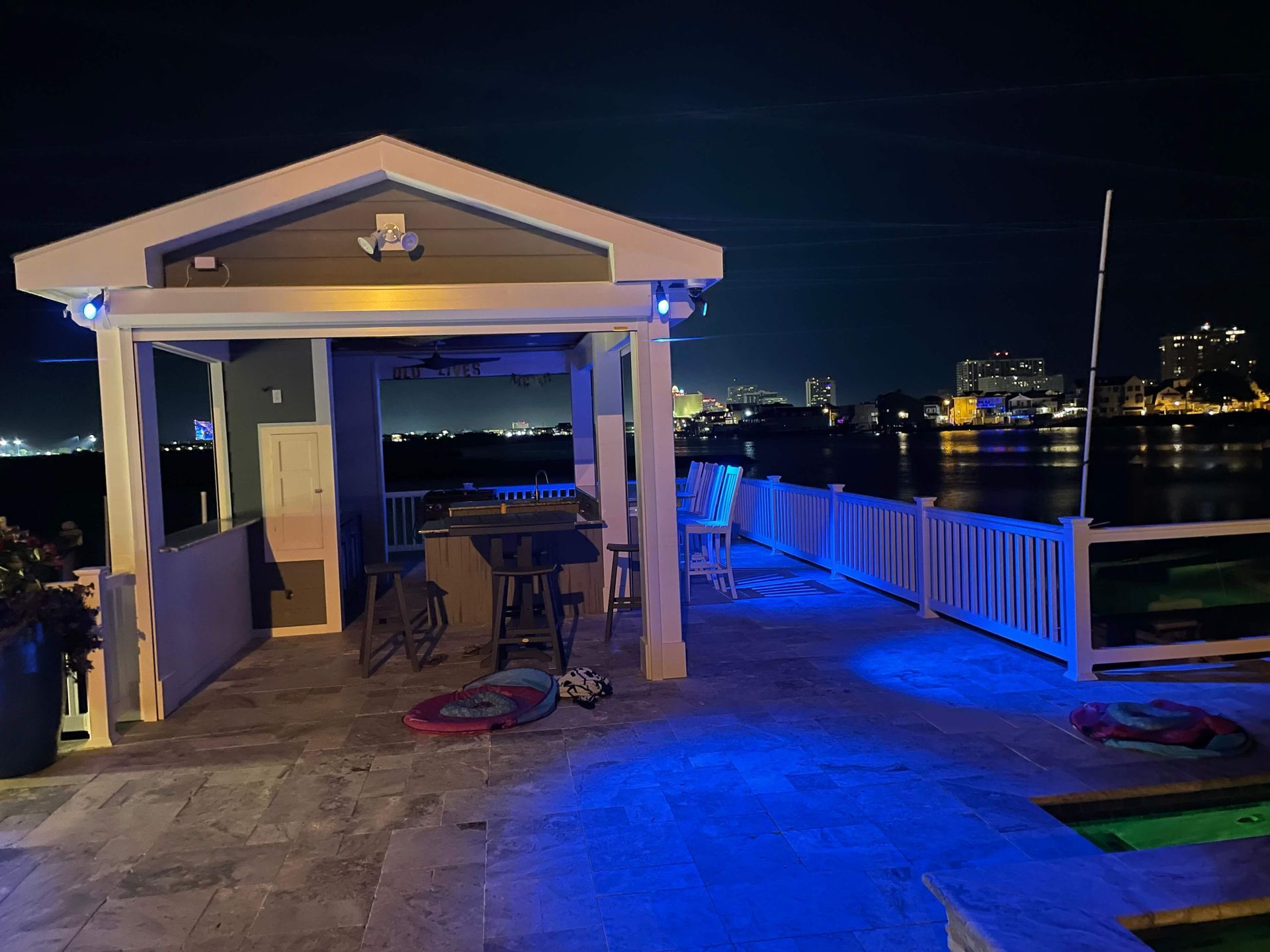 Nighttime waterfront pavilion with blue lighting, a table and chairs, and city lights across the water