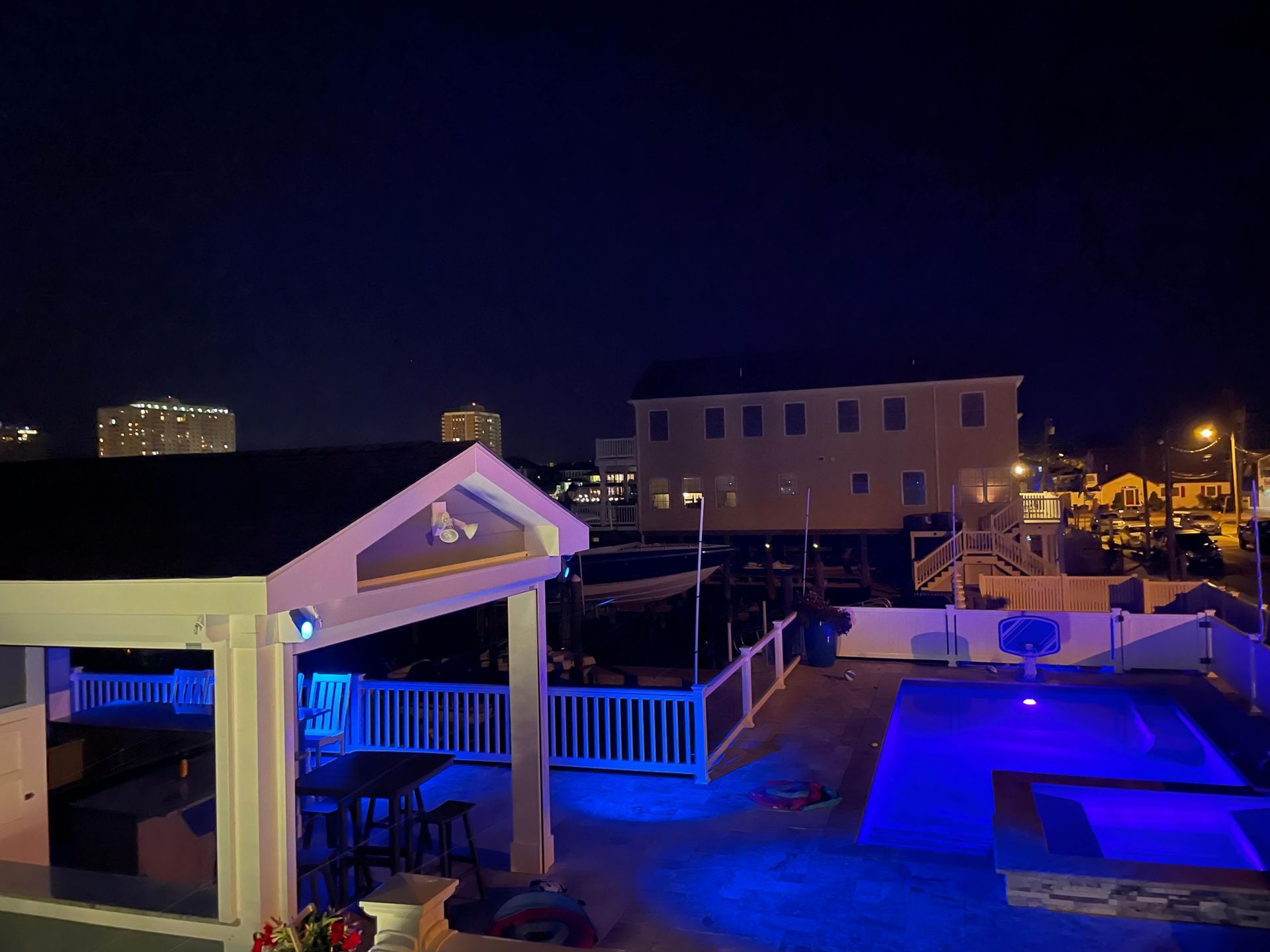 Nighttime rooftop pool deck with blue-lit water, white lounge chairs, and nearby buildings.
