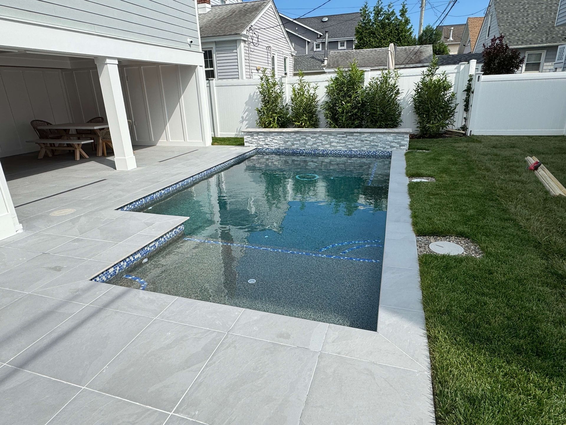 Backyard with a rectangular swimming pool beside a patio and green lawn, bordered by white fencing.