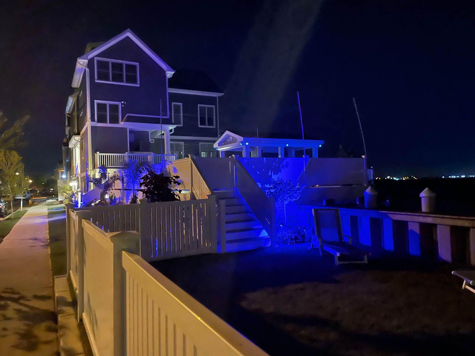 Blue-lit house and deck at night beside a sidewalk and fence