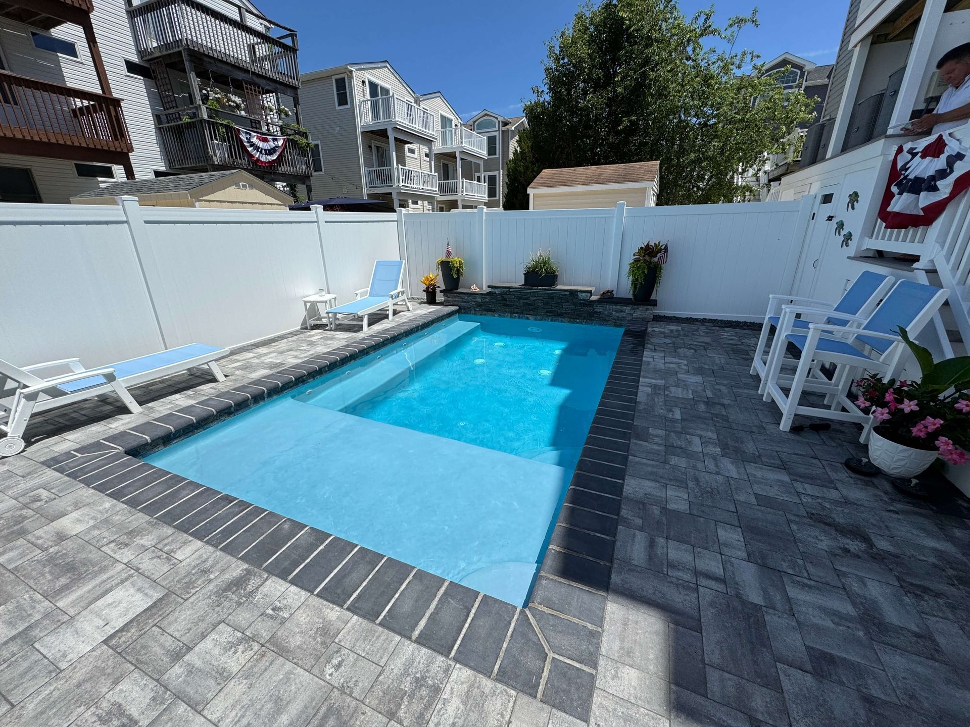 Backyard pool with blue water, gray pavers, white fence, and lounge chairs on a sunny day