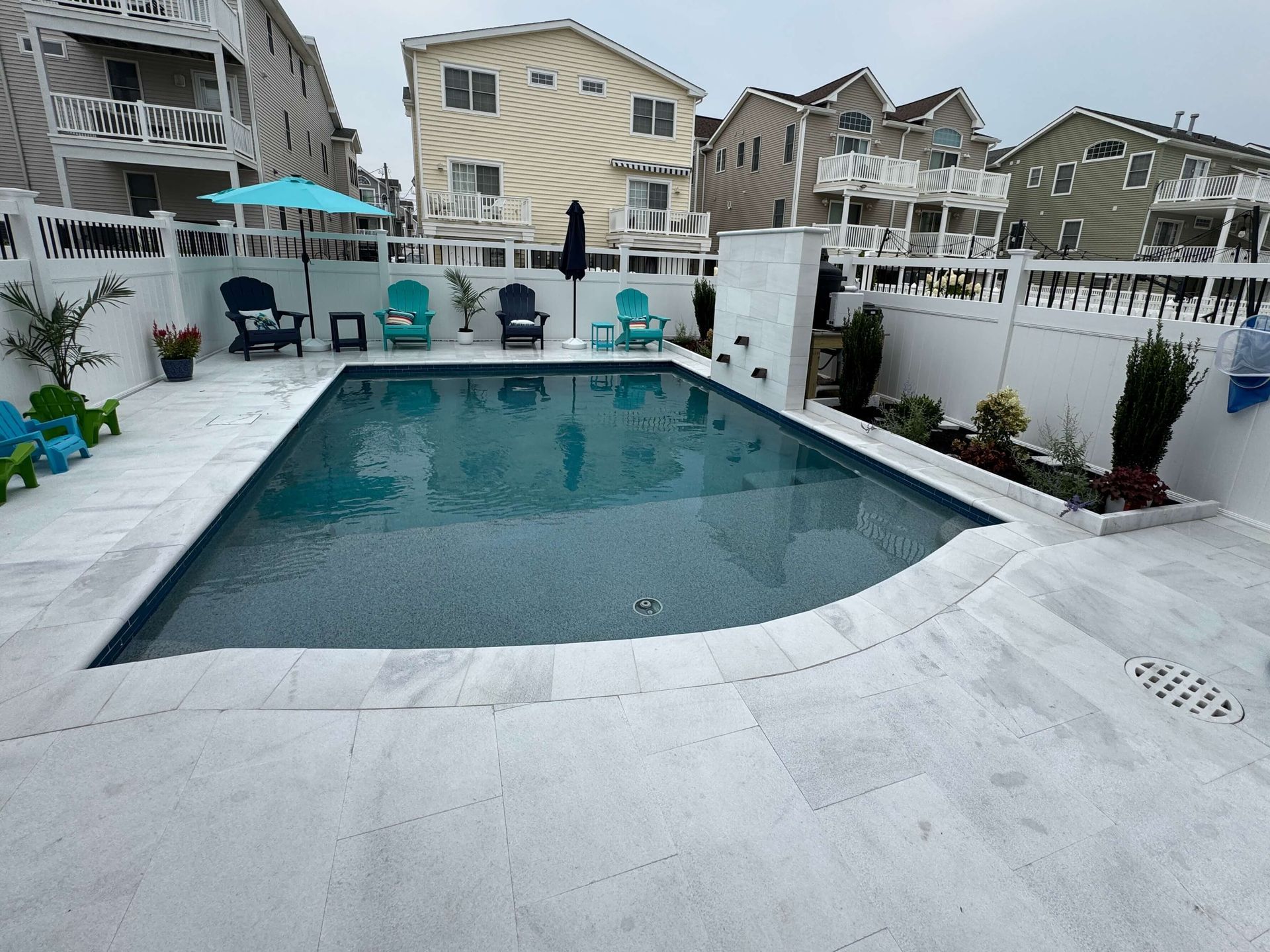 Outdoor backyard pool with white deck, turquoise water, lounge chairs, and neighboring houses in the background