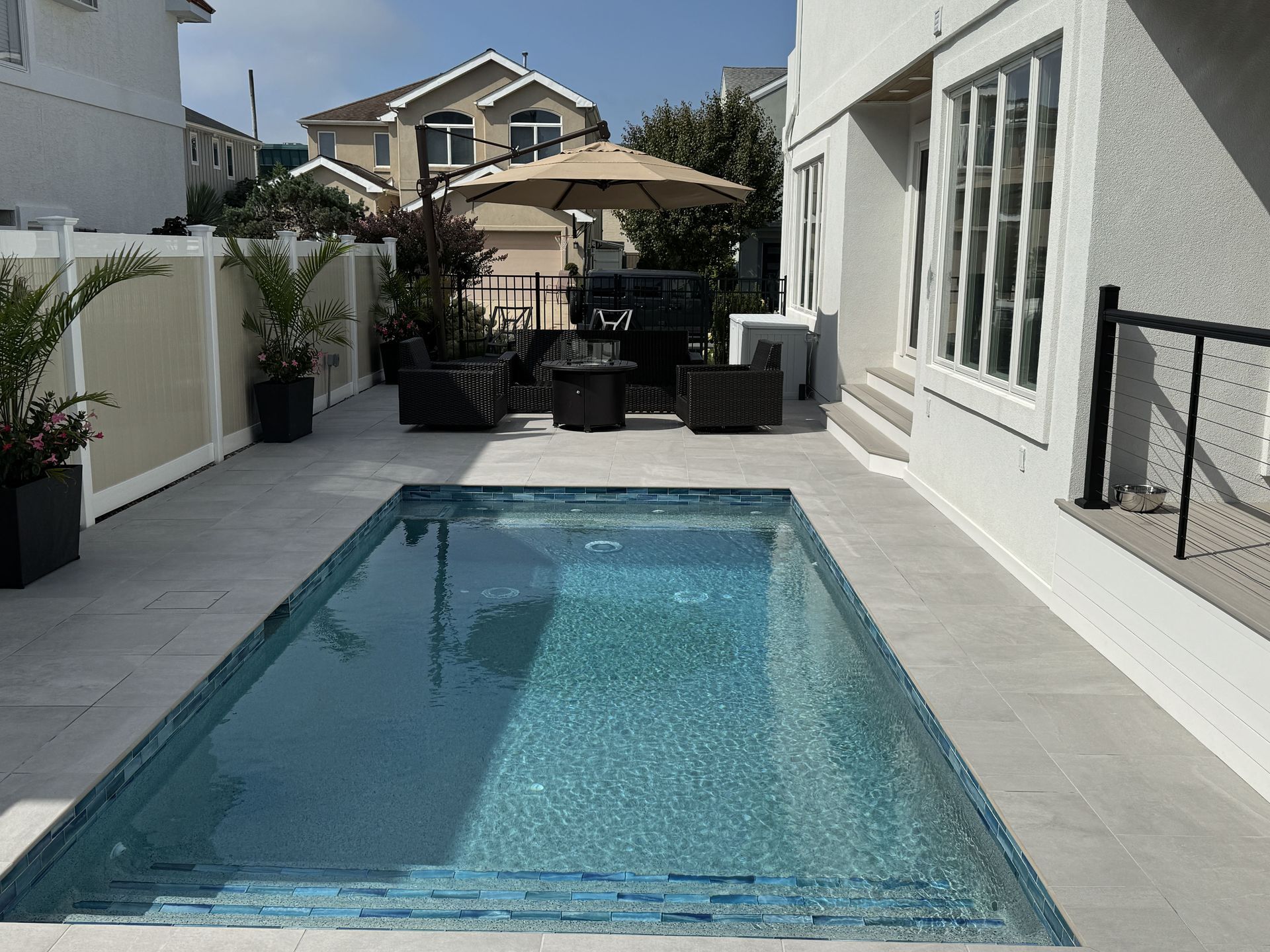 Narrow backyard pool with patio seating, potted plants, and a tan umbrella between white stucco walls.