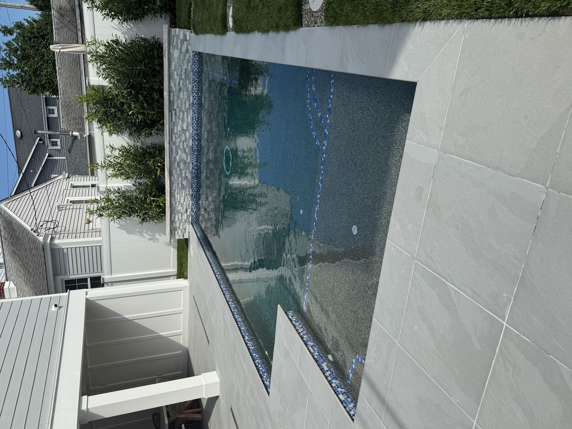 Outdoor pool beside a modern house with white tile and glass fencing, reflecting the sky.