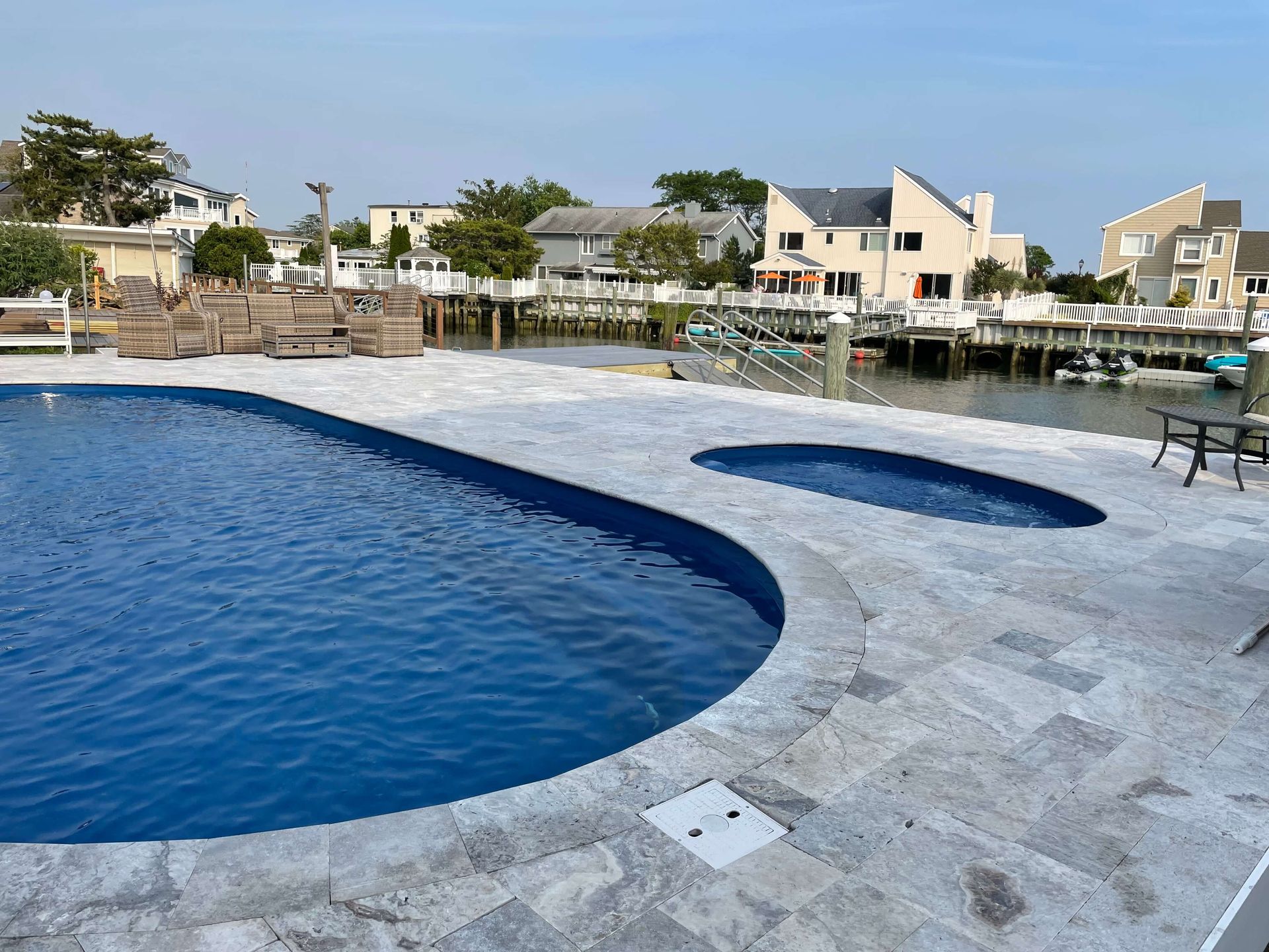 Curved blue pool beside a stone deck, overlooking waterfront homes and boats under a clear sky