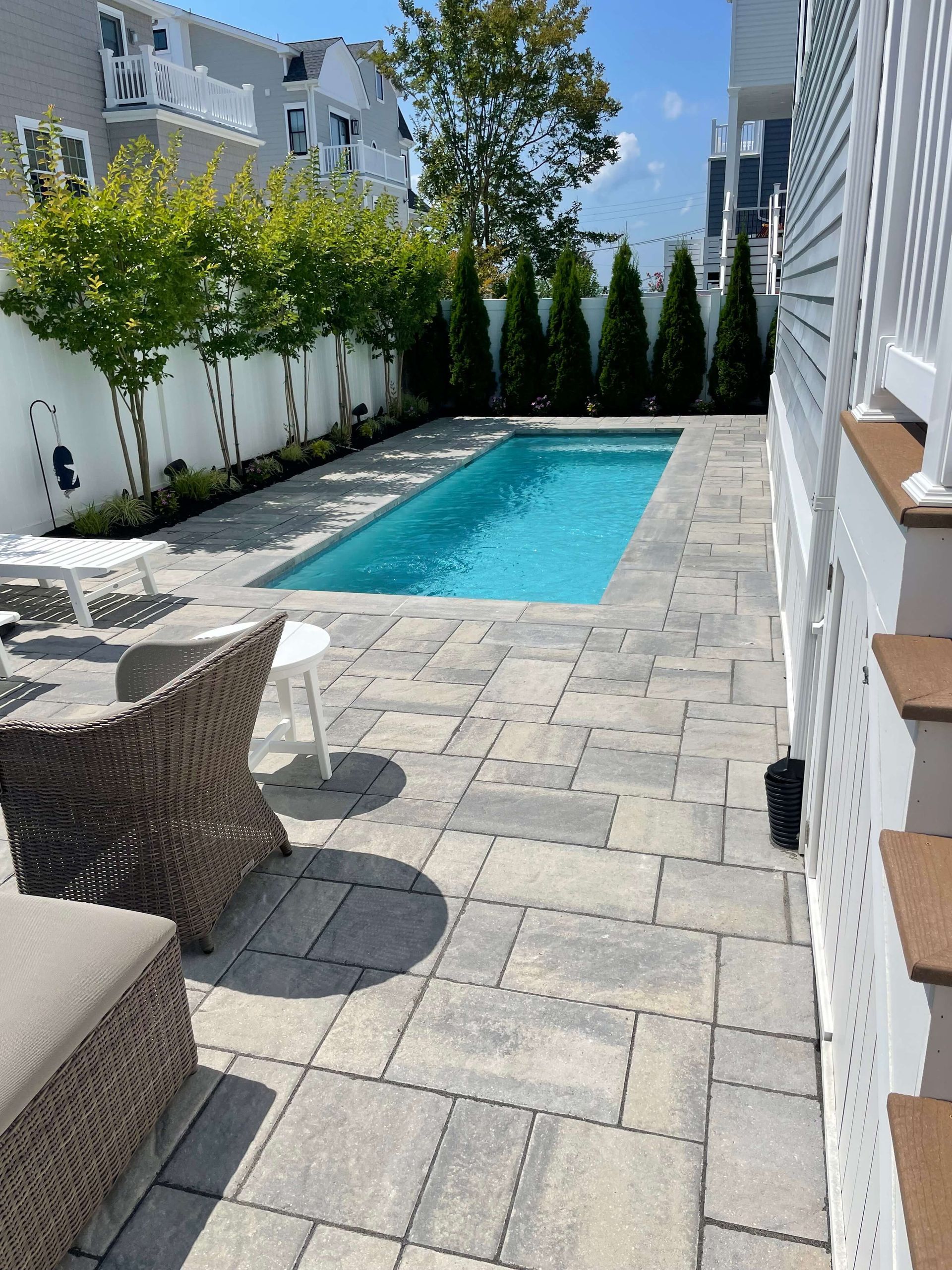 Narrow backyard with a turquoise pool, stone patio, white fence, wicker chair, and tall evergreens