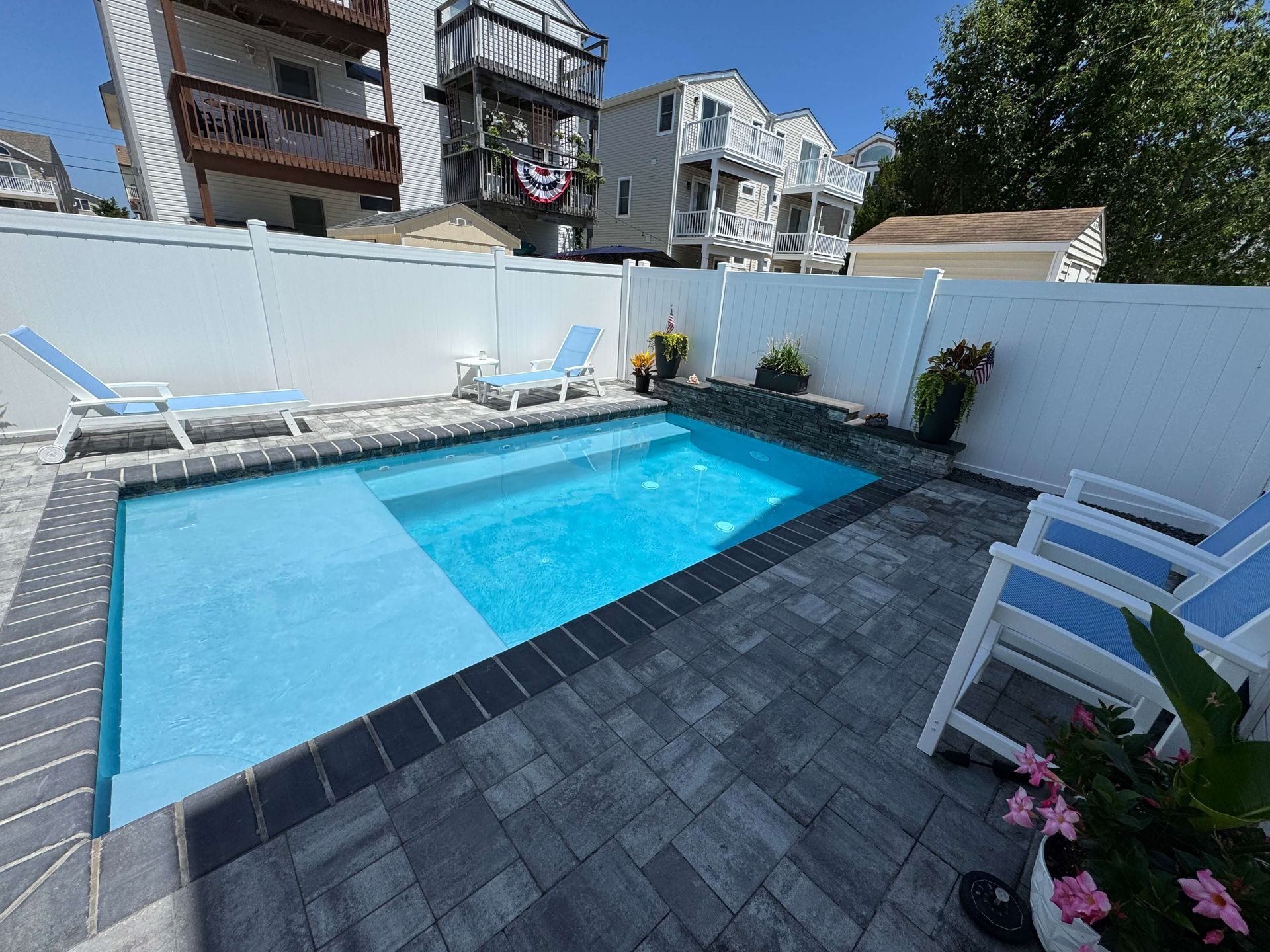 Backyard with a small blue swimming pool, white lounge chairs, and a dark paver deck beside a white fence