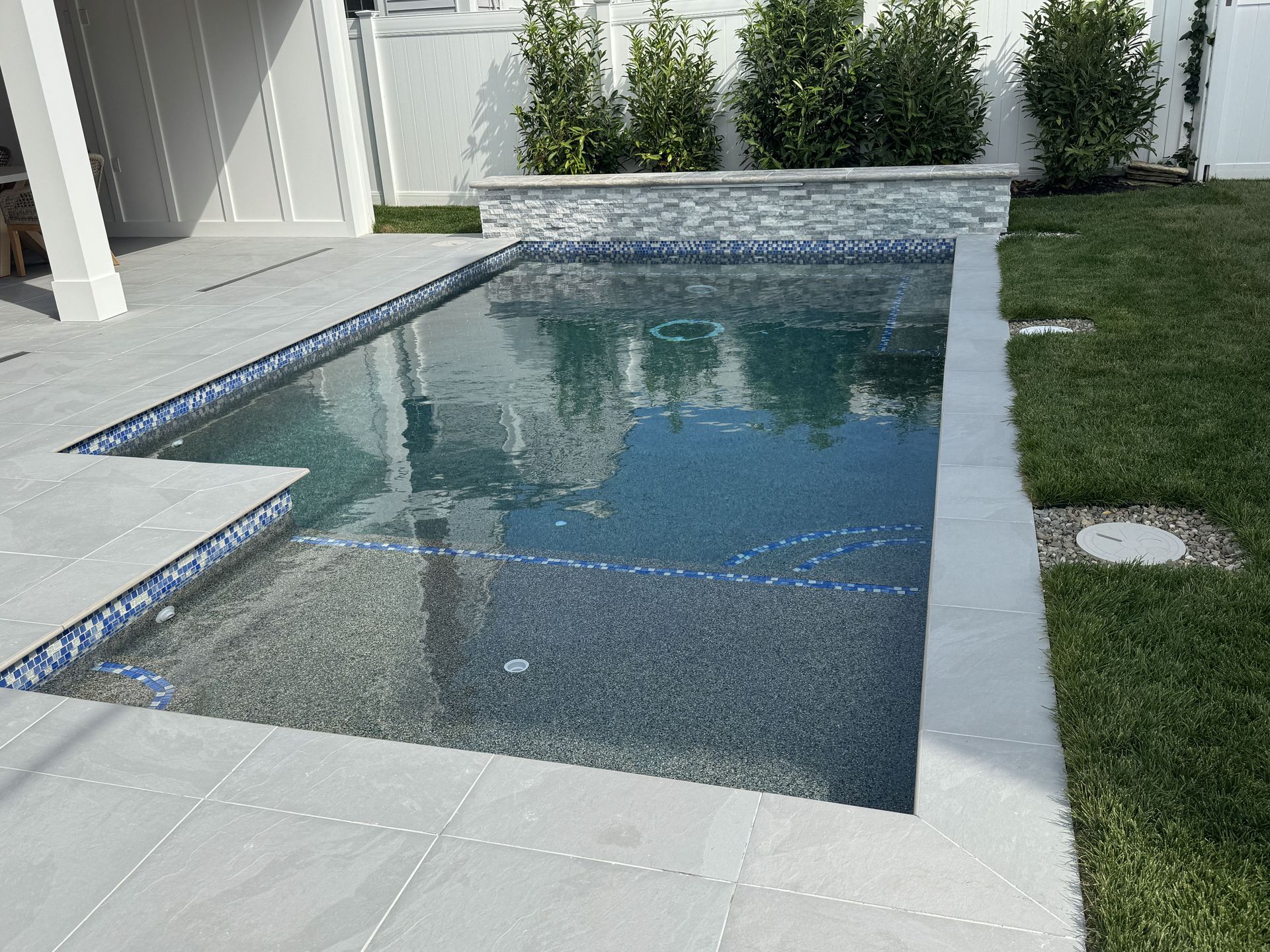 Rectangular backyard pool with blue water, light stone deck, and grass beside a modern house