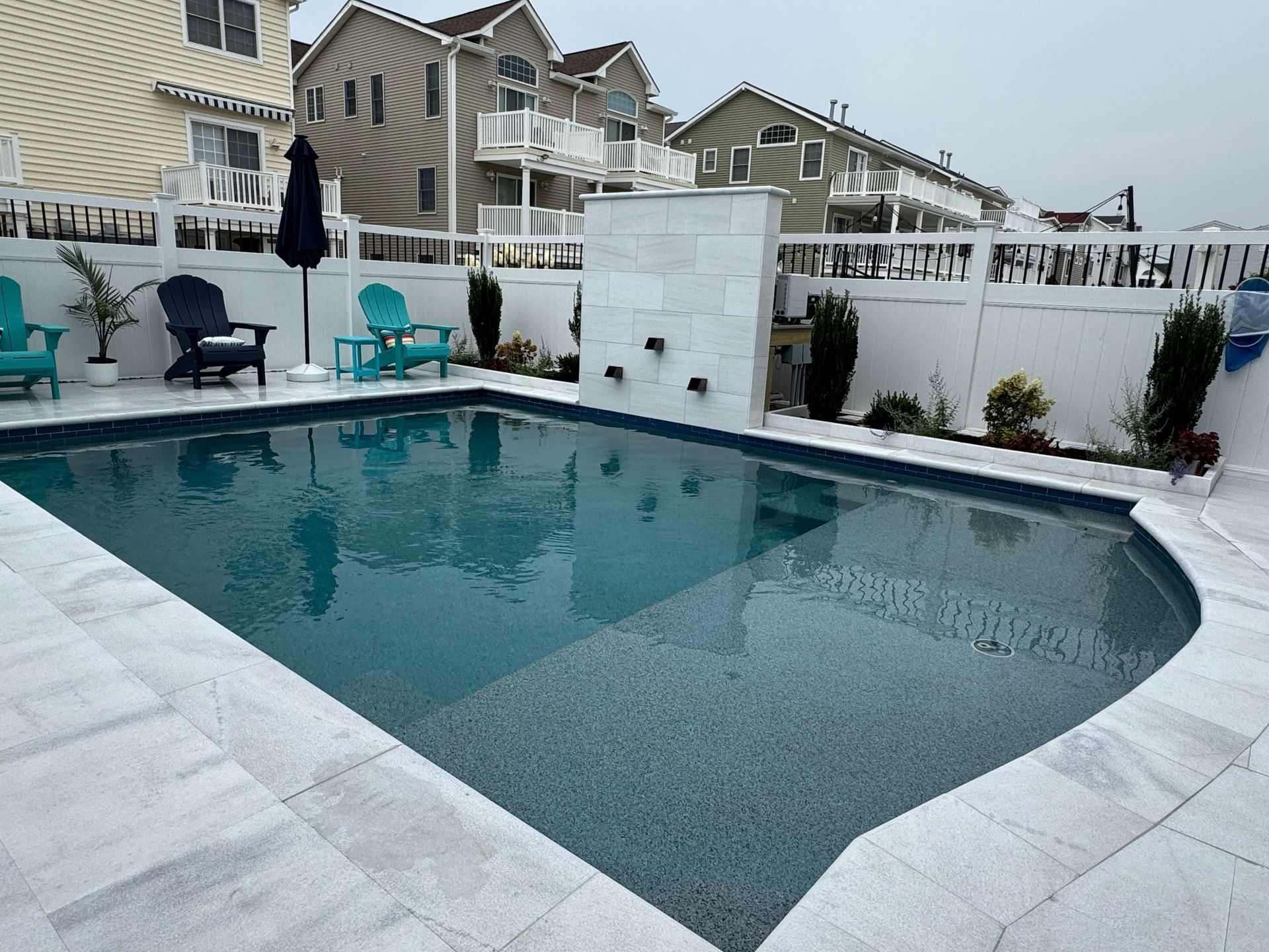 Backyard swimming pool with gray tile deck, white fence, and patio chairs under overcast sky